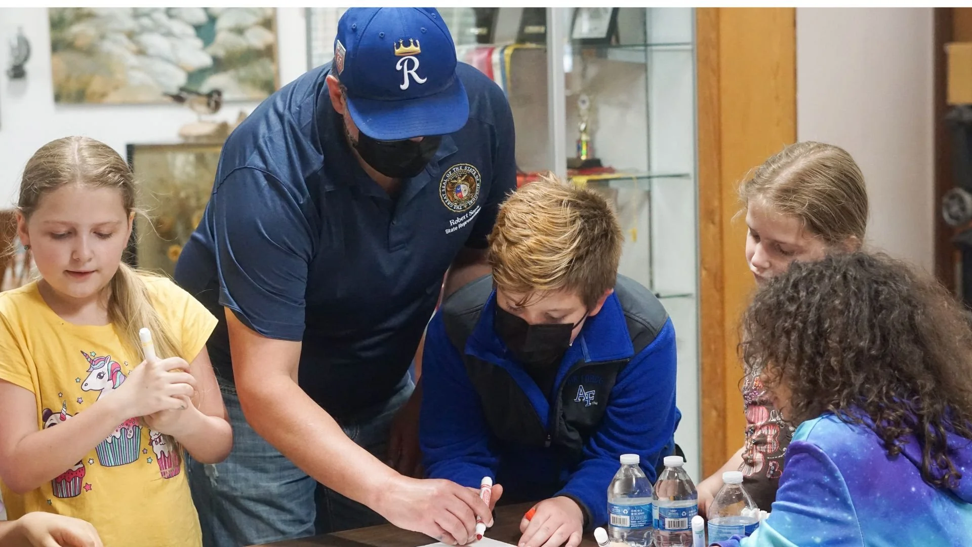 A group of children and an adult man with a badge, all wearing masks, engaged in an activity at a table with water bottles and whiteboards.