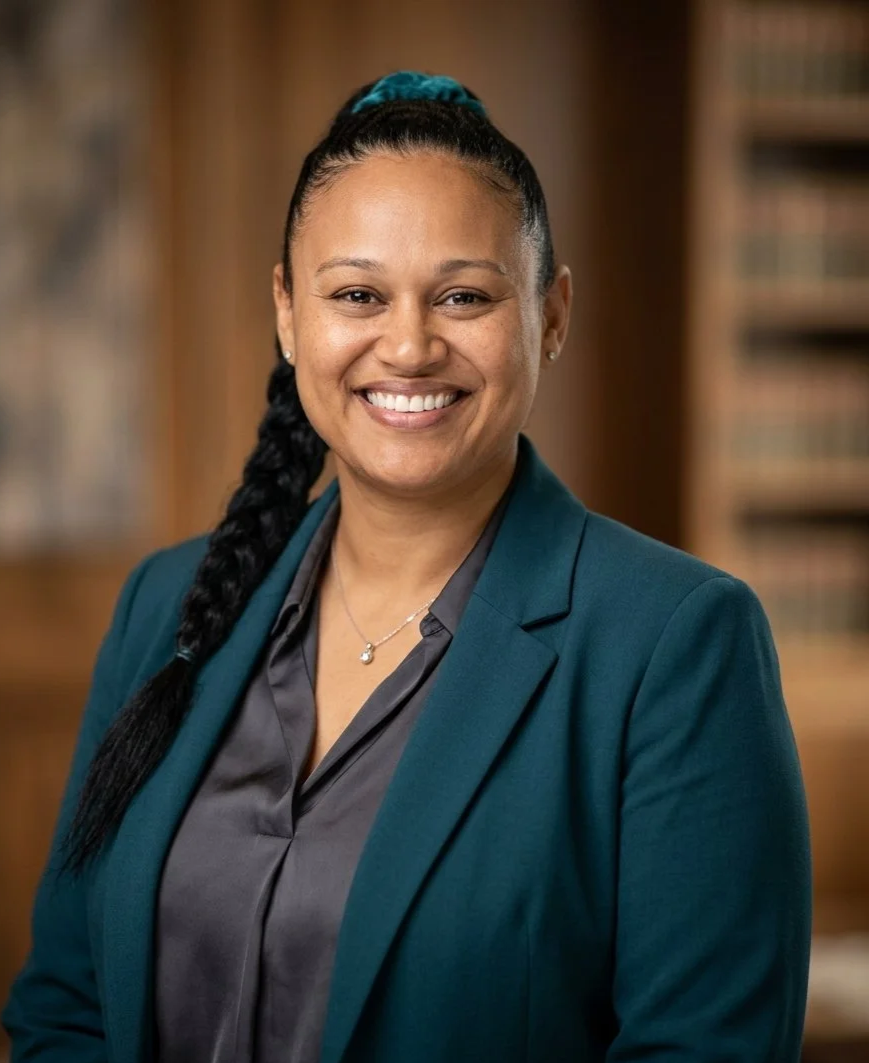 A woman with dark hair in a braid, wearing a teal blazer and a gray blouse, smiling in a library-like setting.