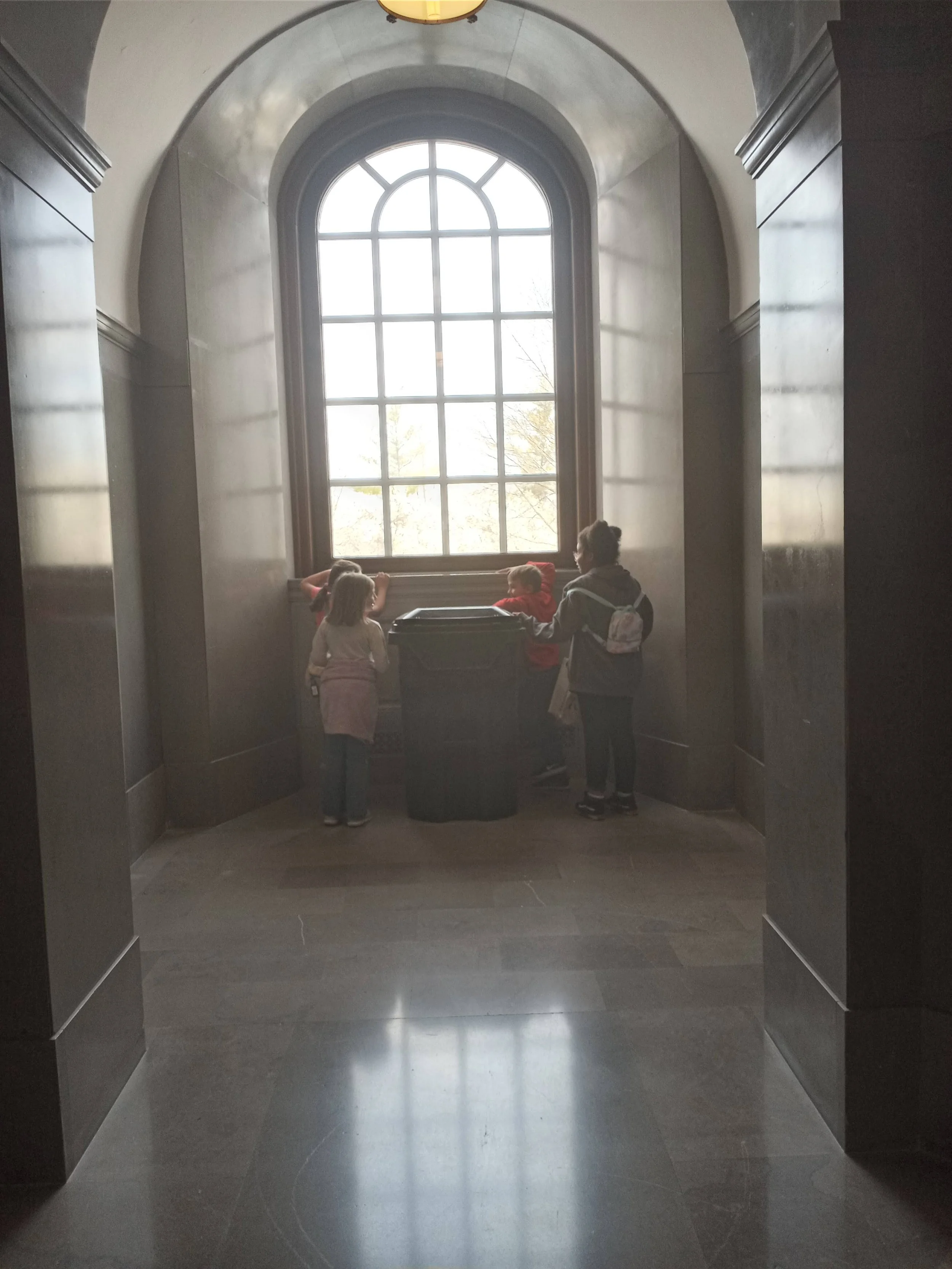 Four children standing in front of a large arched window in a dark hallway, with sunlight shining through the window. Two children are facing the window, one is looking at the camera, and one is facing away.