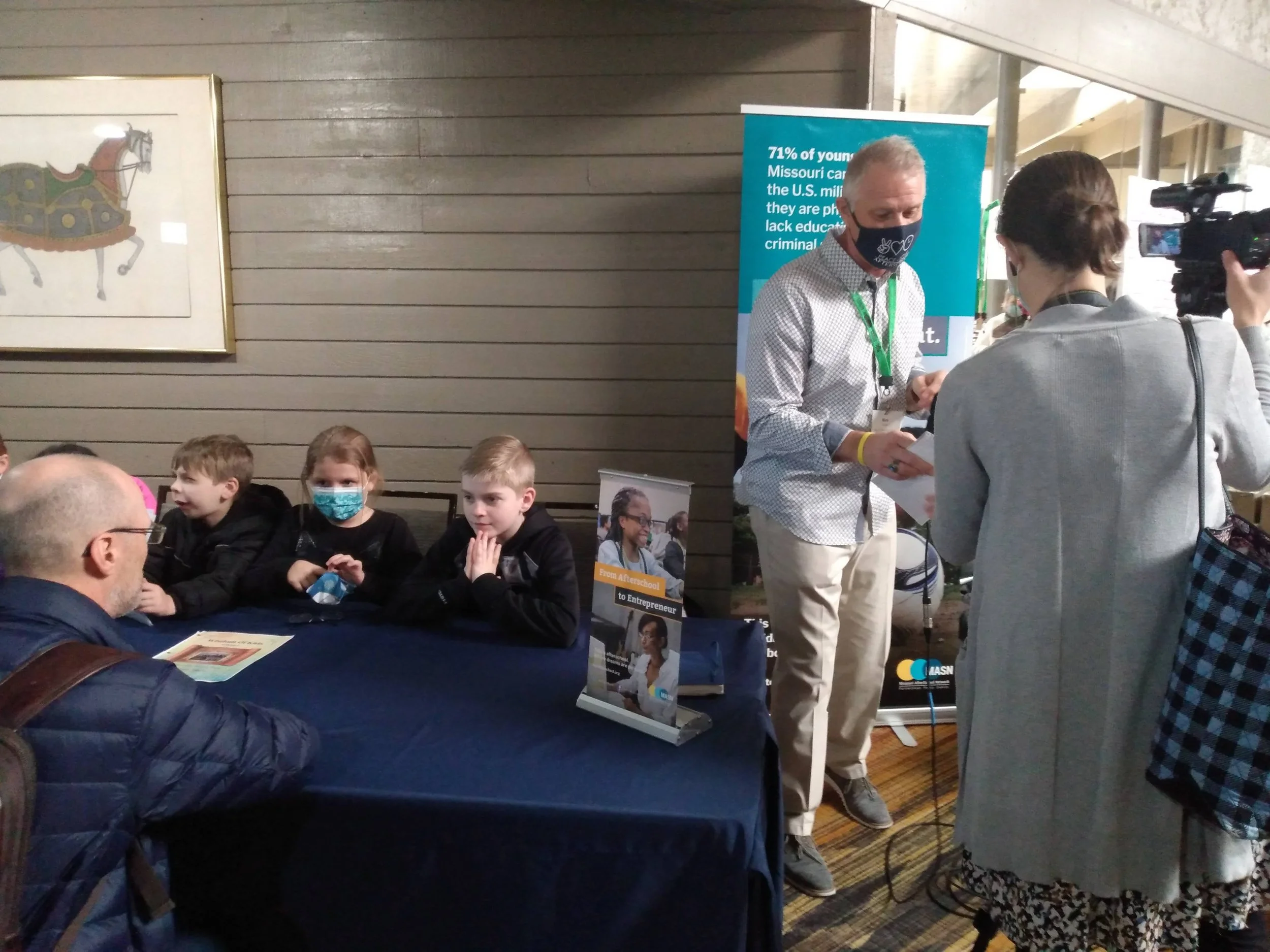 People at a booth talking, with children sitting at a table and a man signing autographs. A woman with a camera films the scene. There are informational posters and banners in the background.