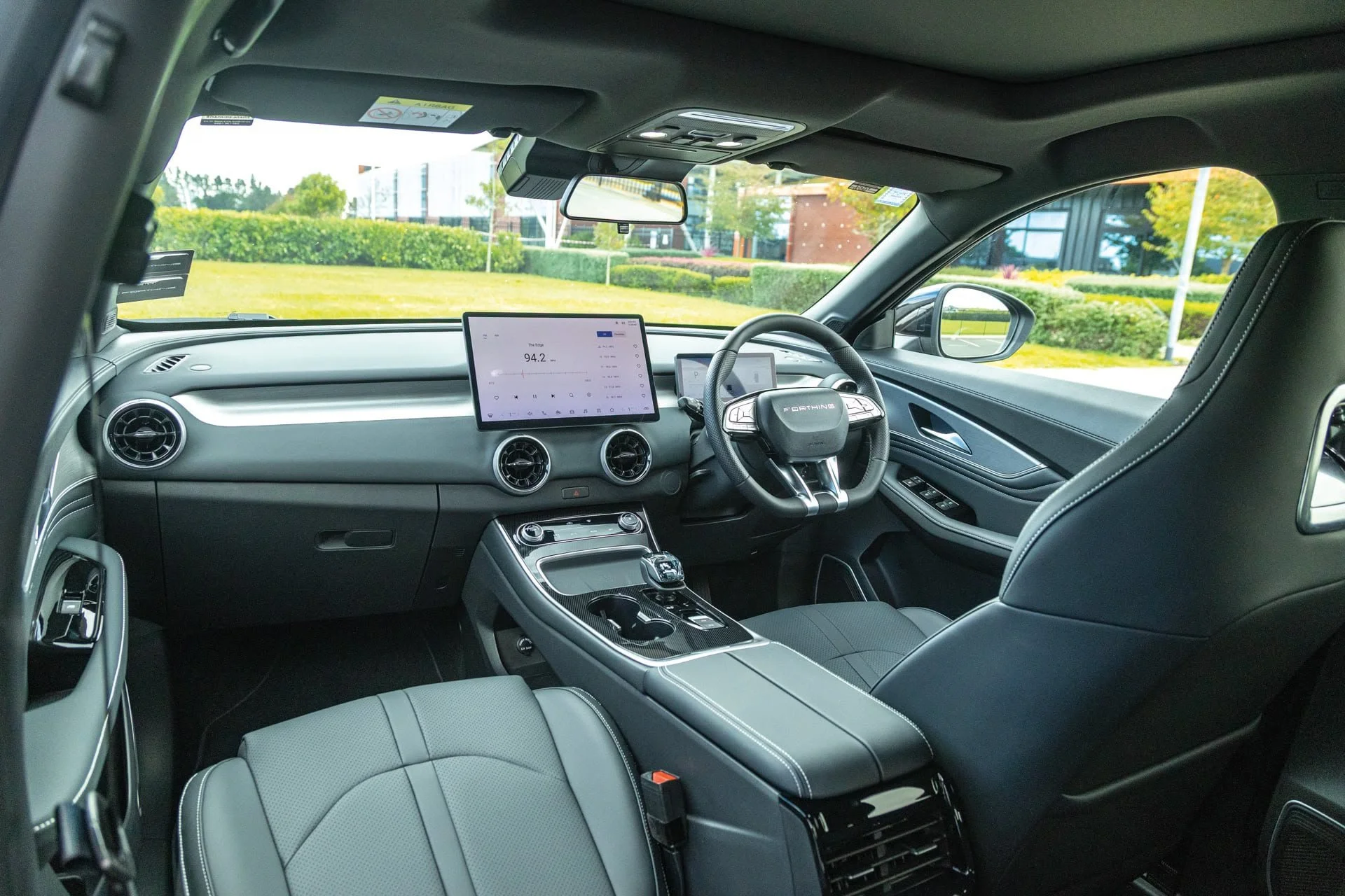 Interior of a modern vehicle with digital displays, steering wheel, and dashboard, parked outdoors near greenery.