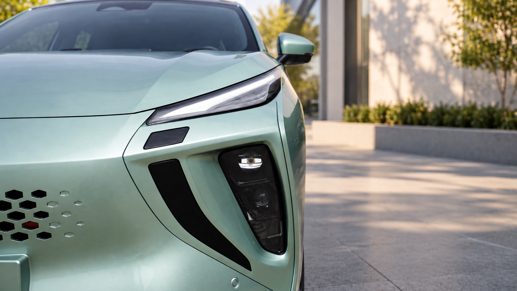 Close-up of the front right side of a modern, mint green electric vehicle parked on a paved area with trees and a building in the background.
