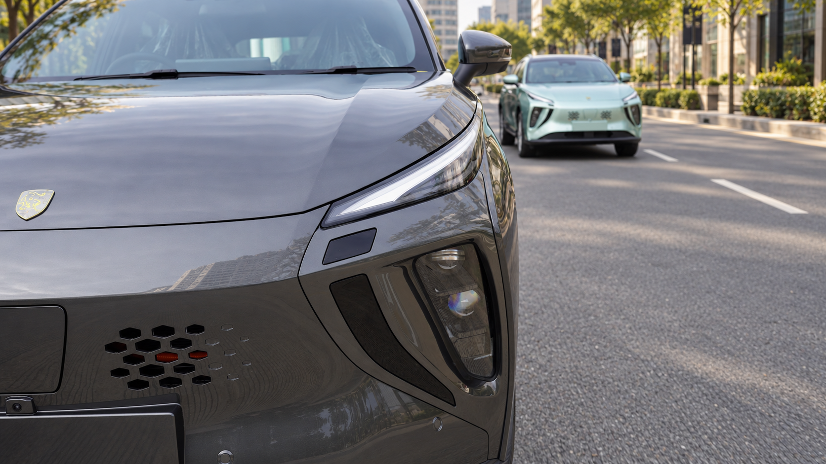 Close-up of the front part of a gray modern electric vehicle with a badge, sleek headlight design, and geometric vents, parked on an urban street with other cars and trees in the background.