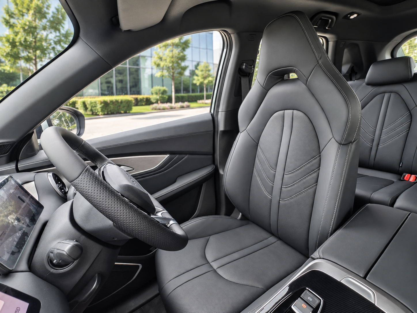 Interior of a modern car showing the driver's seat, steering wheel, and part of the dashboard, with a view of trees and a building outside the window.