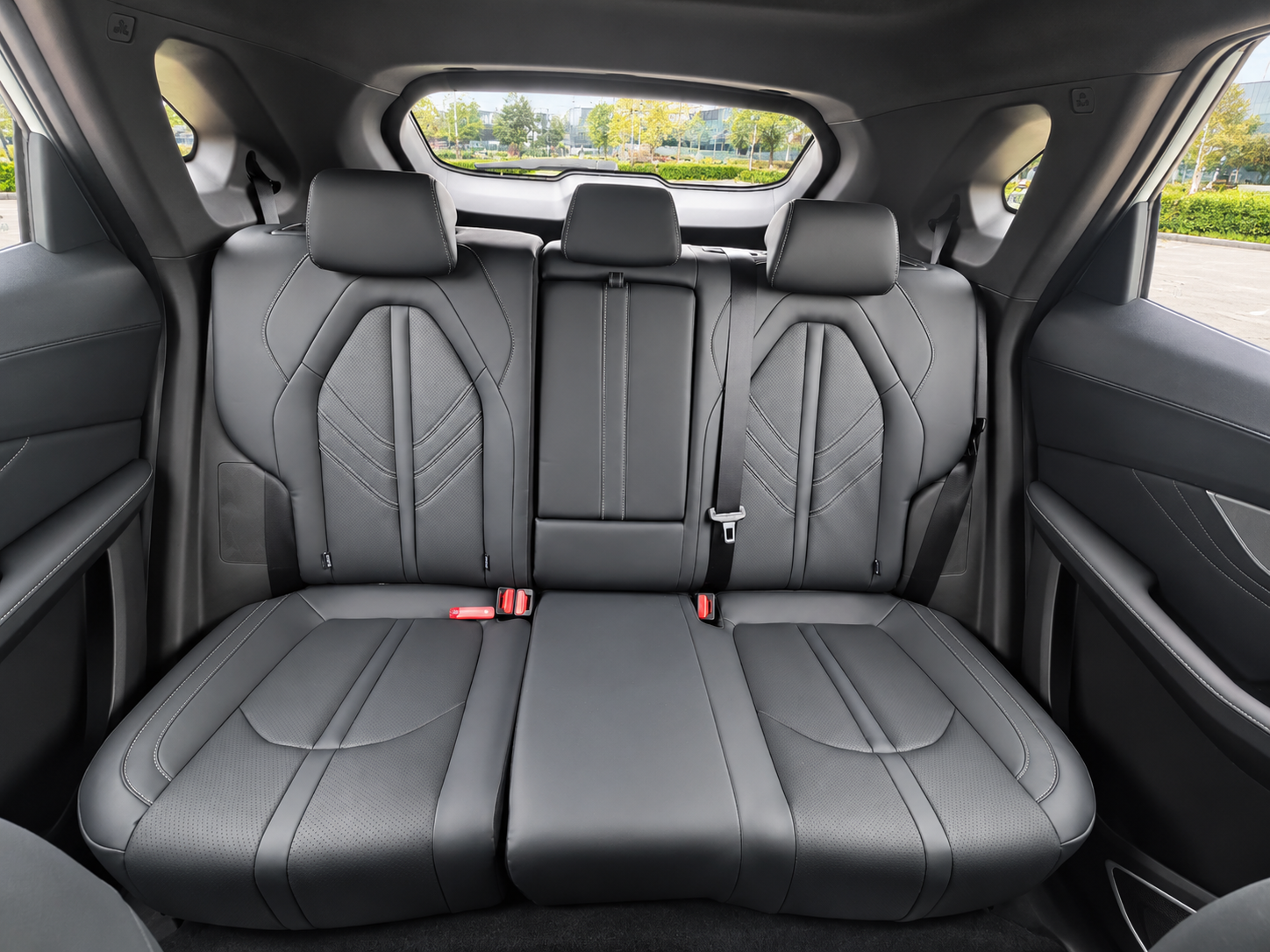 The backseat of a modern SUV vehicle featuring black leather seats with detailed stitching, three headrests, and a view outside through the rear window showing a parking lot and greenery.