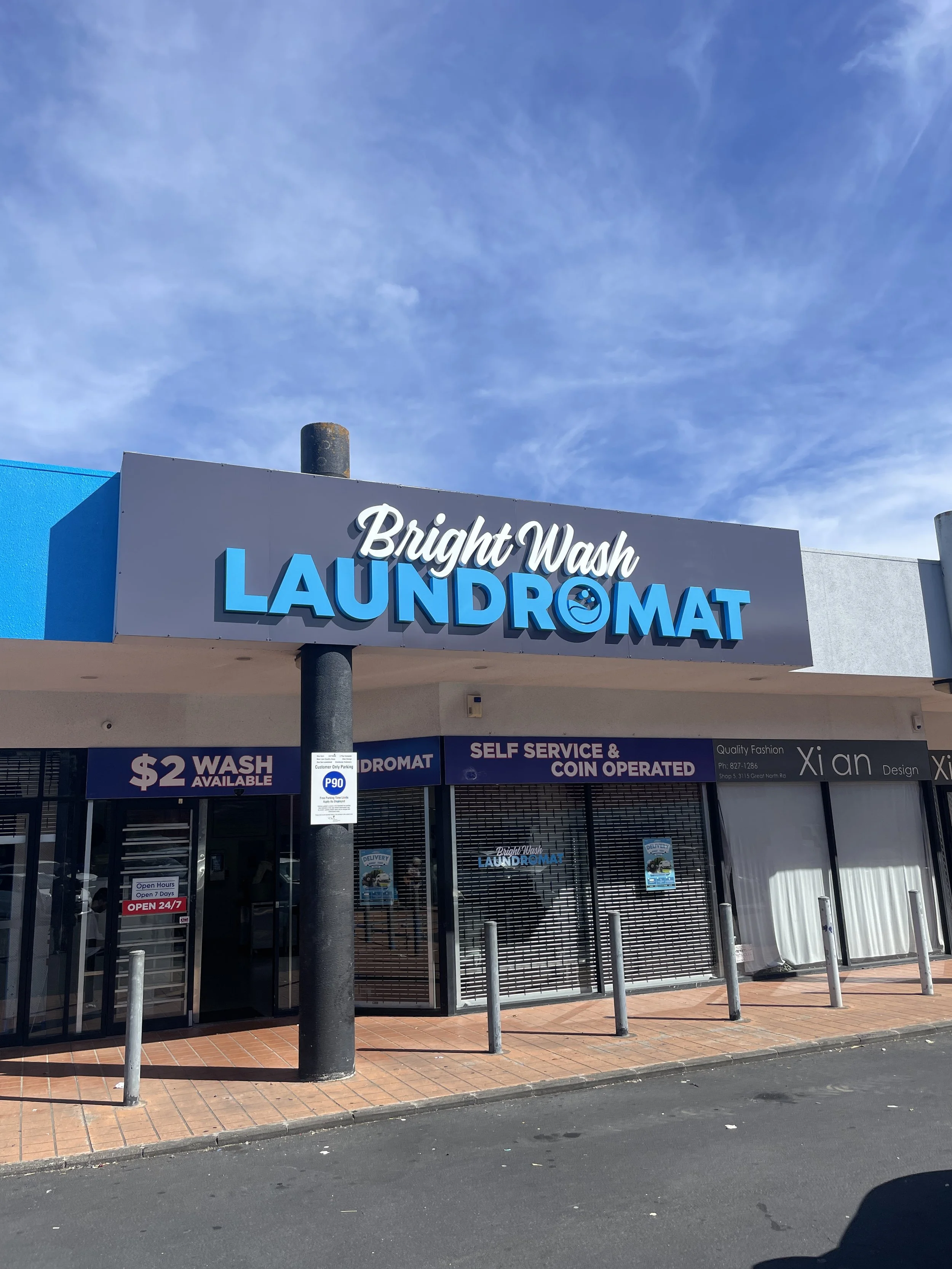 Exterior of a laundromat called Bright Wash Laundromat with a sign indicating $2 wash available, self-service and coin operated. The storefront has closed rolling shutters and a sidewalk with metal bollards.