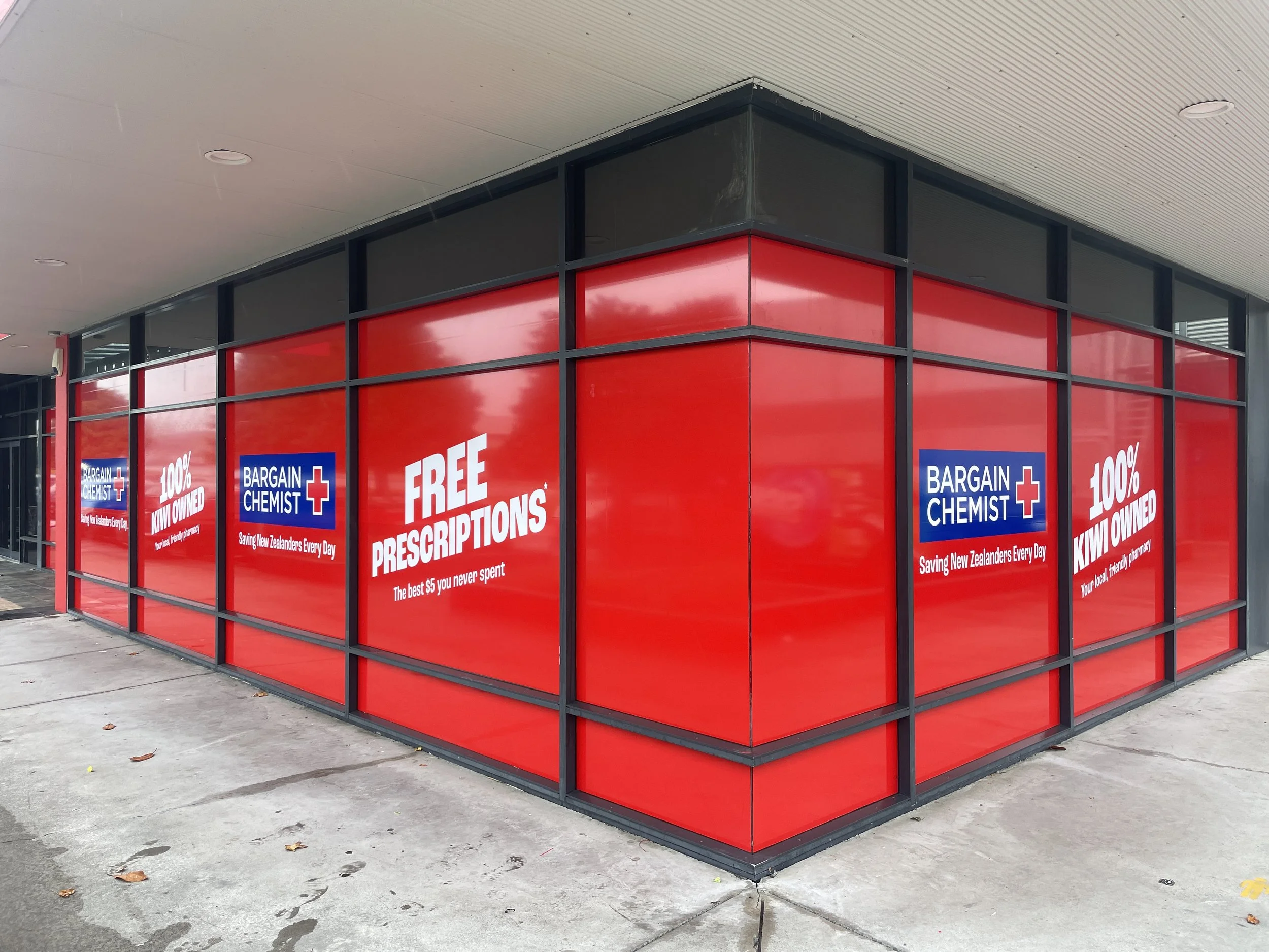 Corner of a building with large red windows and blue signs advertising prescriptions, with the words "FREE PRESCRIPTIONS" and "BARGAIN CHEMIST".