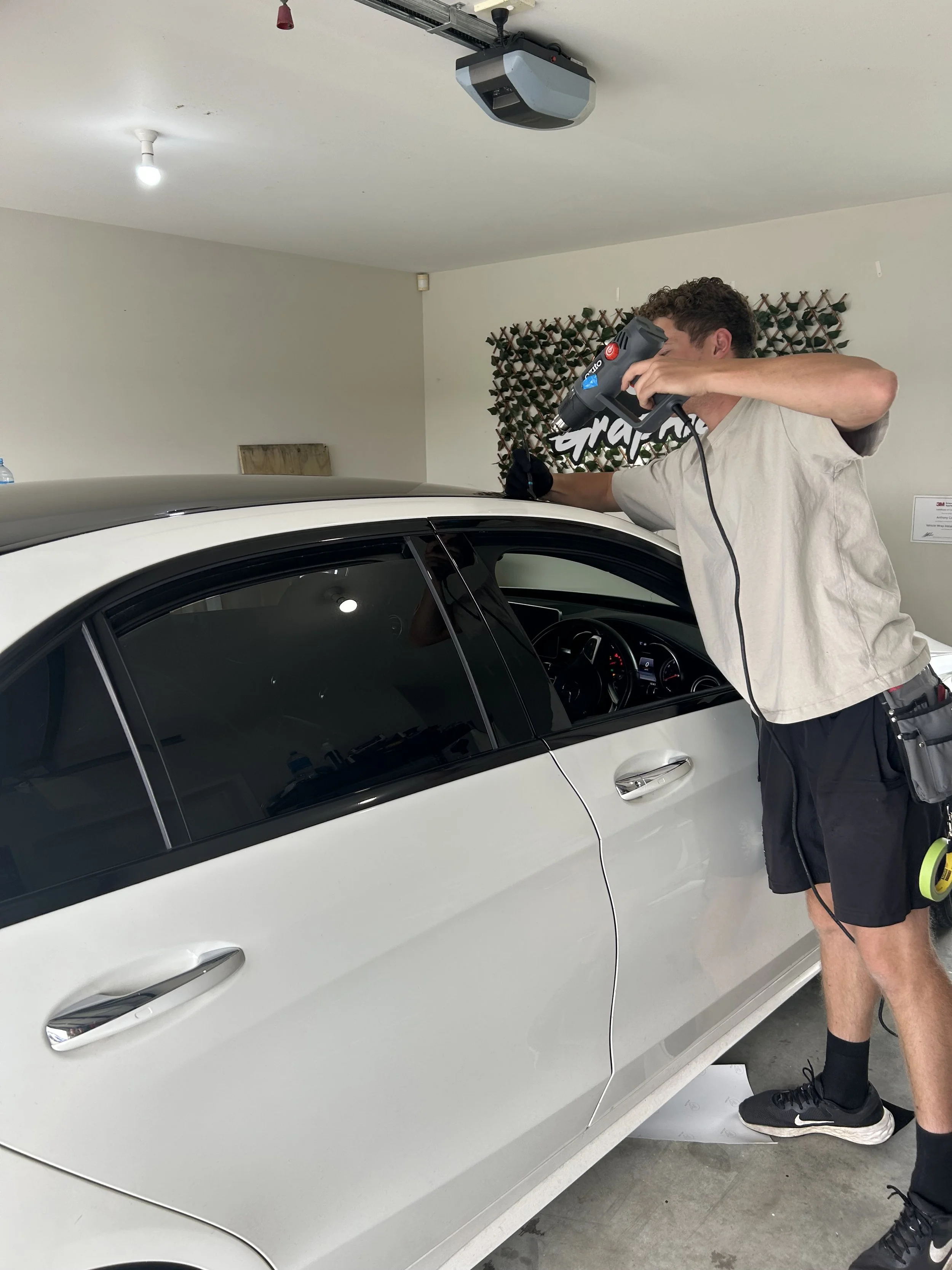 A man is using a handheld device to work on a white car inside a garage or workshop. The man appears to be inspecting or repairing the vehicle.