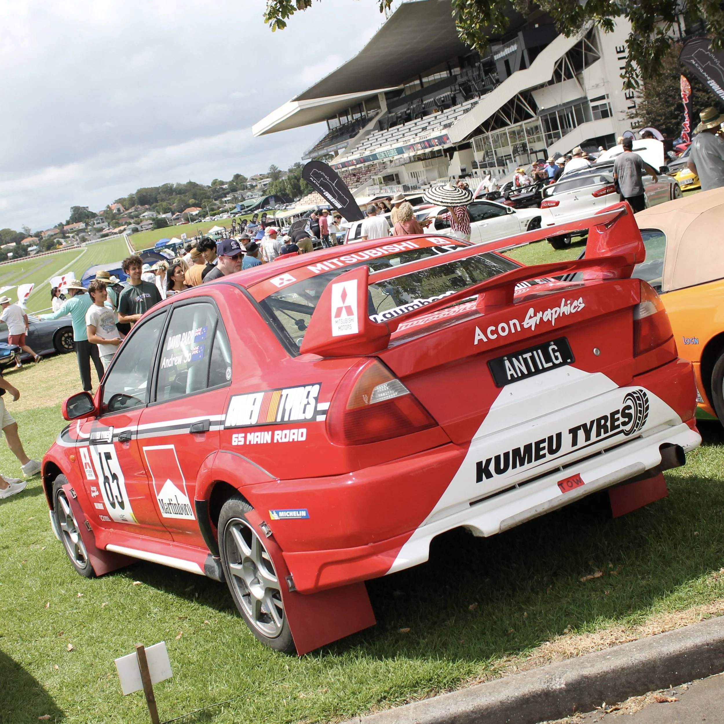 A red racing car with sponsor decals and a large rear wing parked on grass during a motor racing event with a crowd of spectators and a modern building in the background.