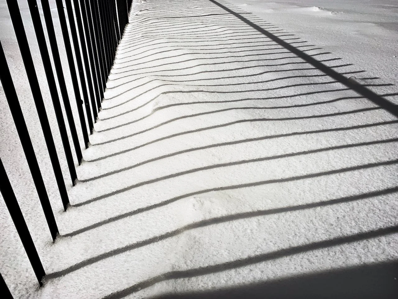 Shadow of a black metal fence creating wavy striped shadow patterns on snow