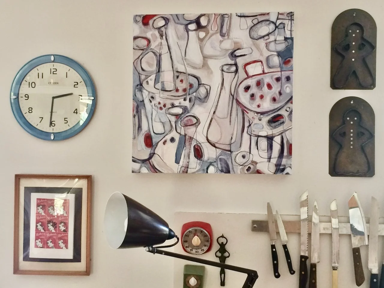 Wall with a blue clock, abstract painting, two black wall decorations, framed art, kitchen timer, bar clamp lamp, small hanging decoration, and various kitchen knives.