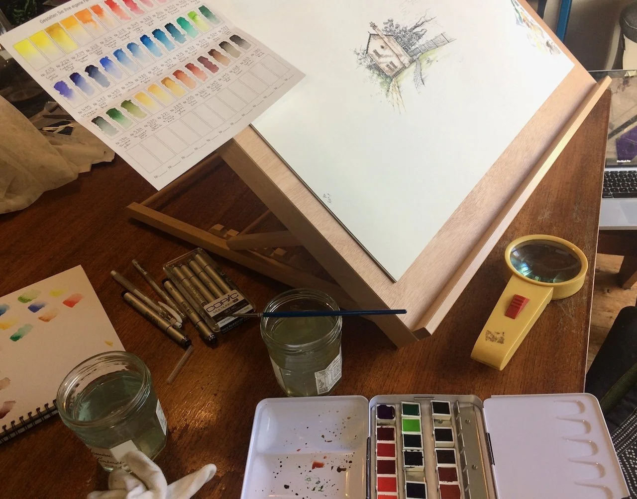 An artist's workspace with watercolor supplies, a sketch of a house on paper, a color palette, water glasses, a magnifying glass, and a laptop on a wooden table.