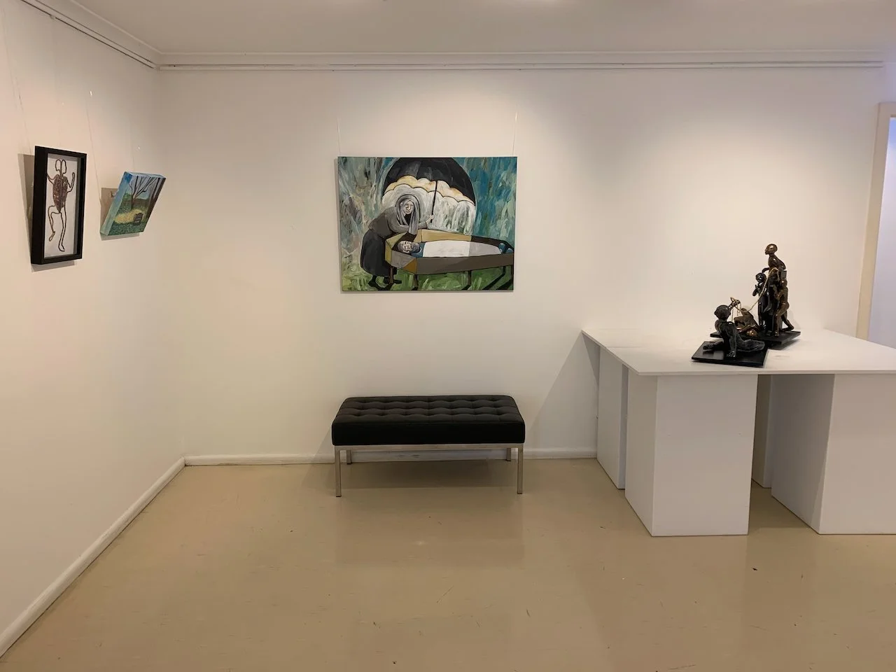 A small art gallery with four paintings on white walls, a black cushioned bench, and a white table displaying black sculptures.