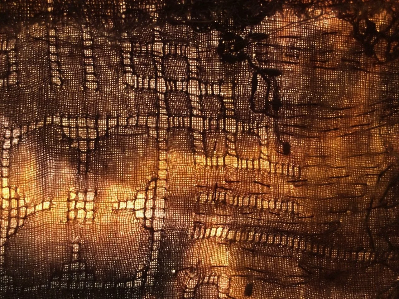 Close-up of a burning candle viewed through a black lace curtain, creating a pattern of light and shadow.