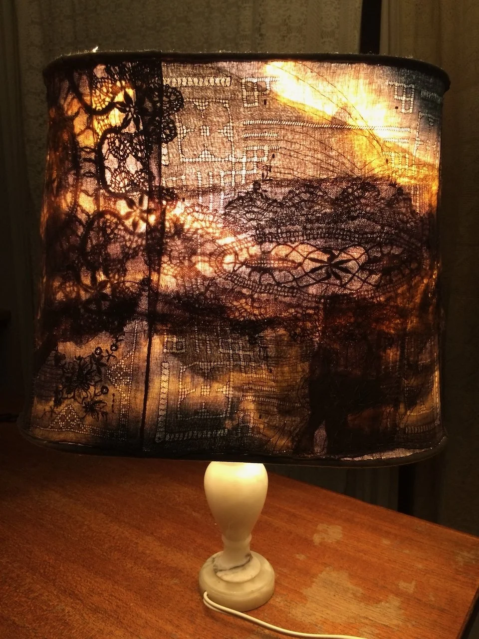 Table lamp with a lace-patterned fabric lampshade emitting warm light, placed on a wooden surface.