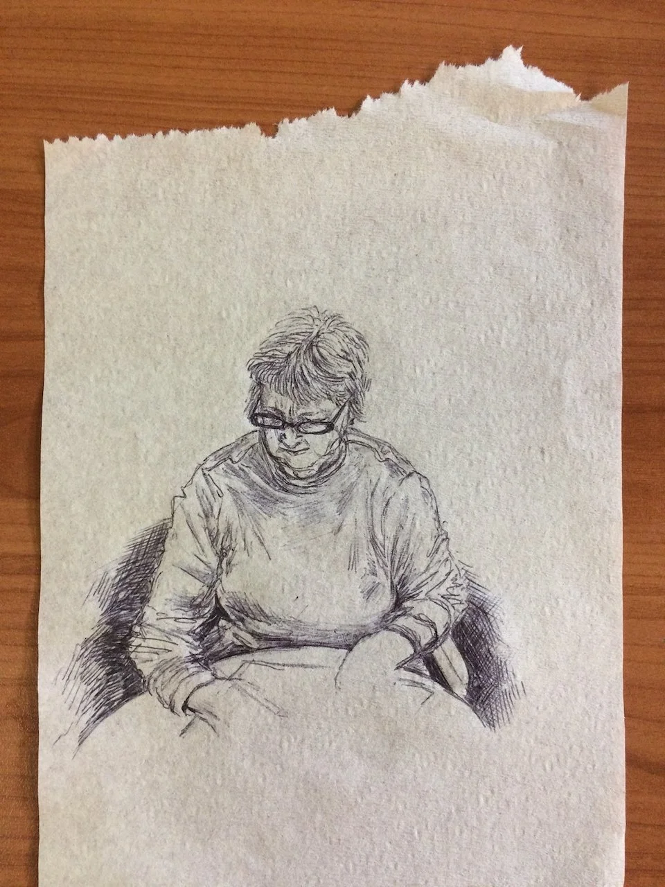 A pencil sketch of a person wearing glasses, sitting with hands in lap, wearing a long-sleeved shirt, on a torn piece of paper placed on a wooden surface.