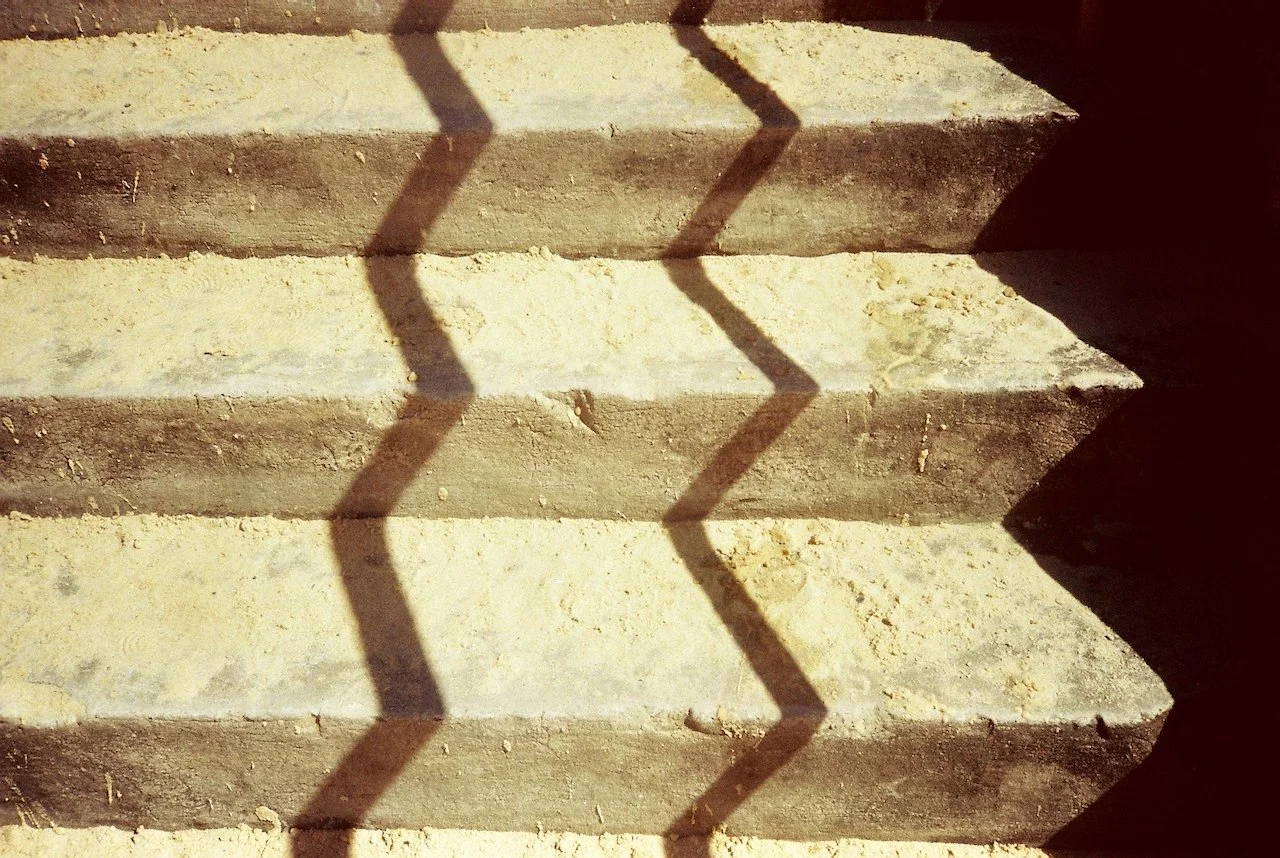 Concrete stairs with a diamond-shaped patterned shadow cast on them, indicating sunlight from the side.