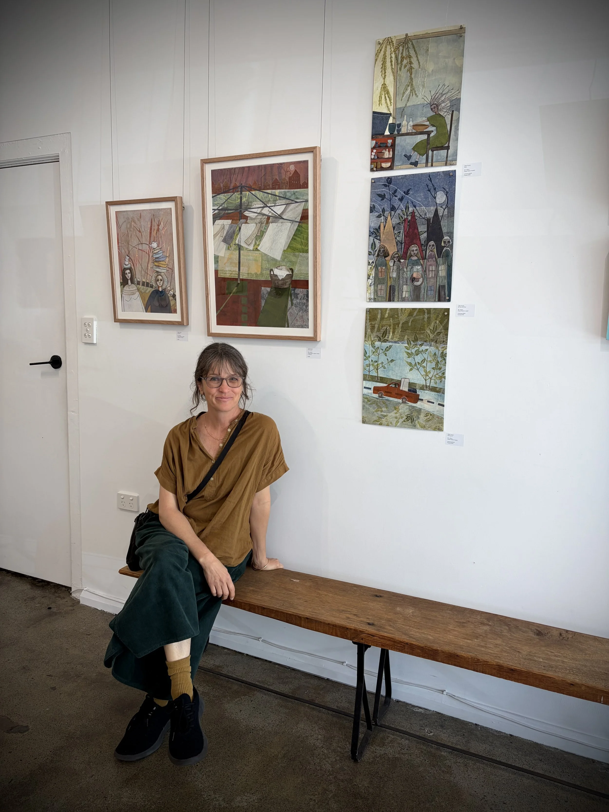 A woman with glasses and brown hair is sitting on a wooden bench in an art gallery. Behind her, a white wall displays five colorful paintings arranged vertically. The woman is wearing a brown shirt, dark green pants, mustard-colored socks, and black sneakers. She has a black bag strap across her chest.