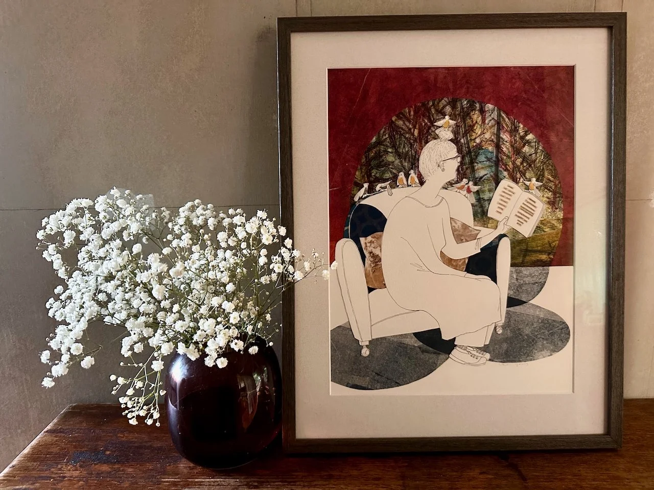 A framed illustration of a person reading a book, seated on a chair in a room with a large window showing a forest scene with birds, and a vase of white flowers on a wooden surface next to the frame.