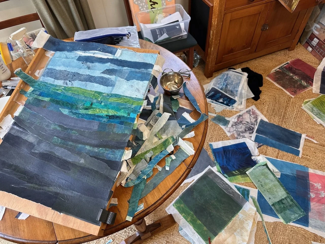 A table covered with torn and cut pieces of artwork, mostly in blue and green hues, some organized on a wooden frame and others scattered on the table and floor. Various art supplies are also visible in the background.