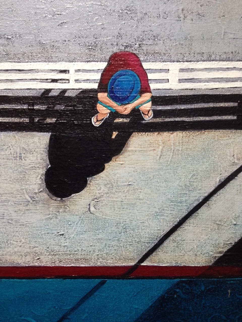 Top-down view of a person sitting on a bench, wearing a red shirt, black pants, and a bright blue hat, with their shadow cast on the ground.