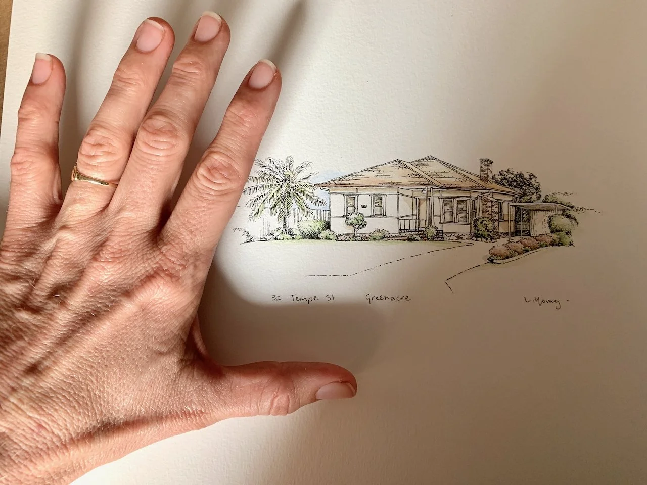 A hand with a wedding ring is placed over a sketch of a house. The house is a single-story structure with a front porch, situated in a landscaped yard with trees, shrubs, and a palm tree.