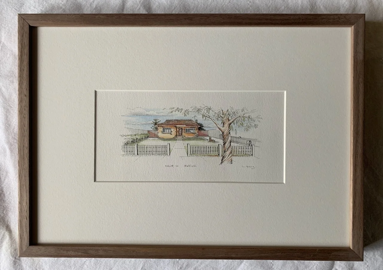 A framed watercolor drawing of a house with a fence and a large tree in front of it, under a blue sky with clouds.