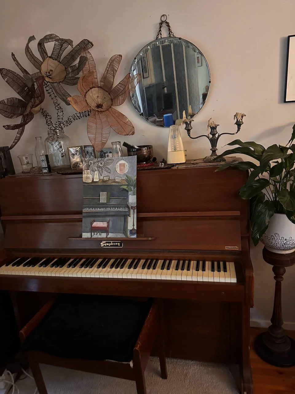 A wooden upright piano with a painting on top, decorated with various small objects. To the right, a tall plant in a white pot, with wall decor including a circular mirror, a candelabra, and large decorative flowers made of woven material on the wall