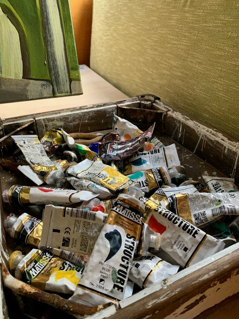 A wooden box filled with used acrylic paint tubes, some crumpled and stained, with a paint palette in the background.