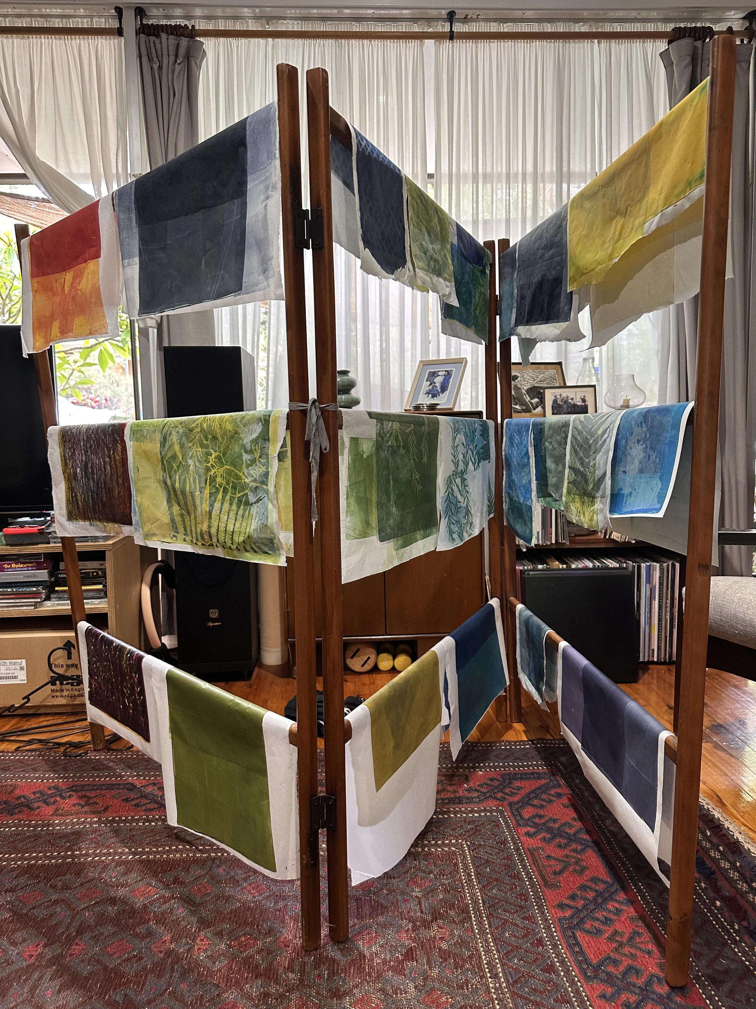 A wooden room divider with fabric artwork hanging on it, set up inside a room with large windows covered by white curtains, a bookshelf with framed photos, and a patterned area rug.