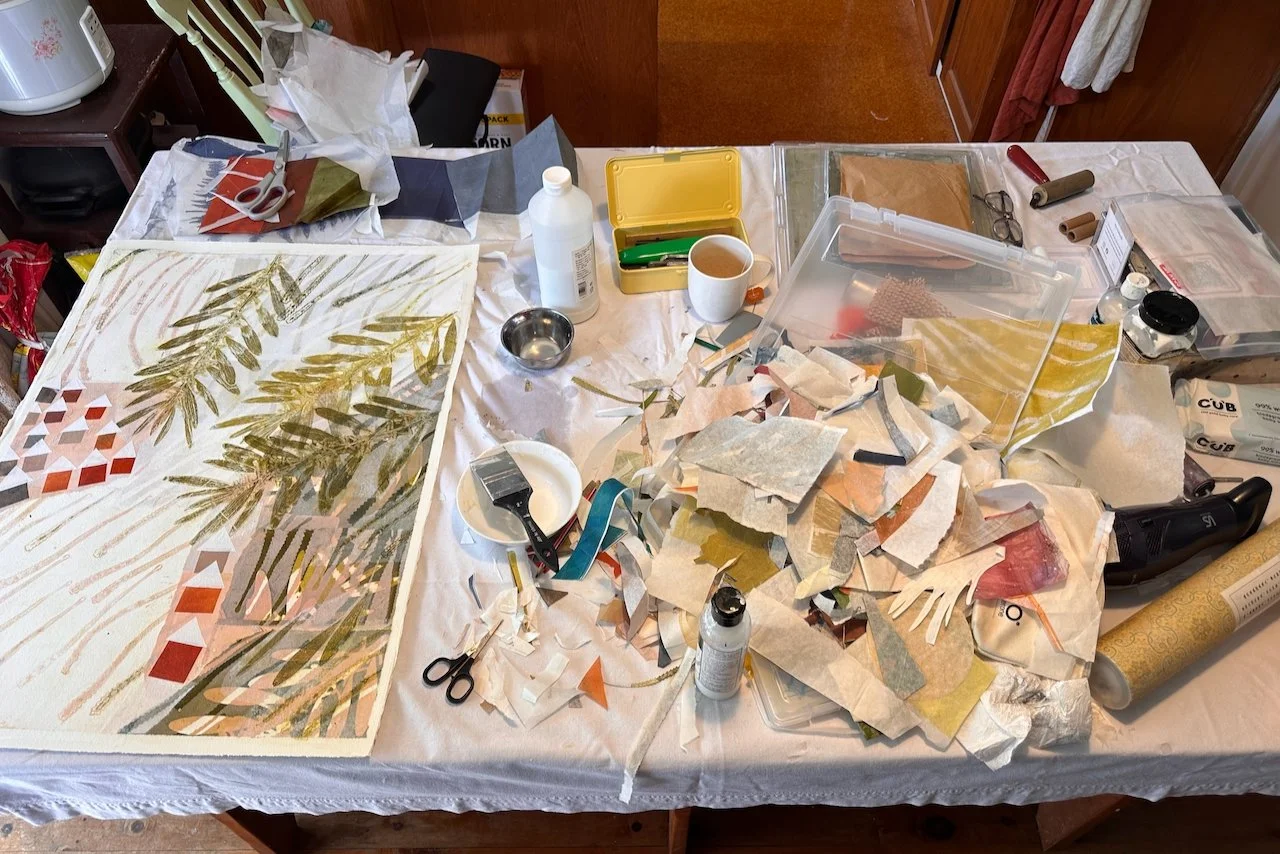 A cluttered workspace with art supplies and paper, including a partially finished botanical painting of green leaves, scissors, paintbrushes, paints, and various cut-out papers and scraps.