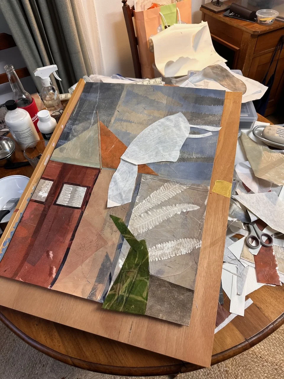 The image shows a craft project on a wooden table, consisting of various pressed leaves and botanical specimens arranged on a glass or acrylic sheet with a wooden frame. Surrounding the project are craft supplies including scissors, glue, paper, and 