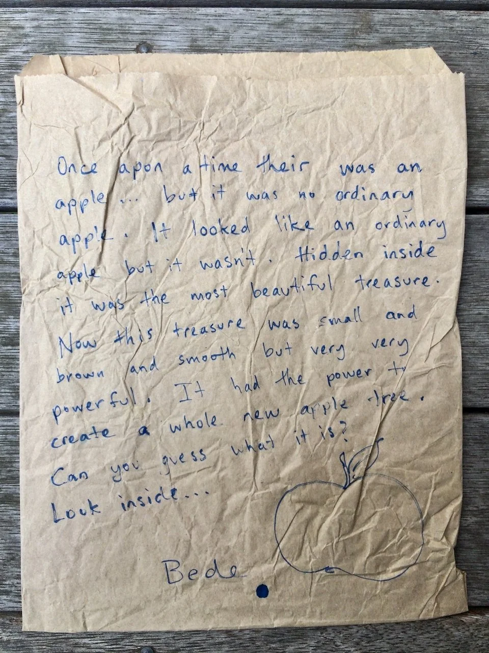 A handwritten note on crumpled paper with a drawing of an apple and a small apple illustration at the bottom, laying on a wooden surface.