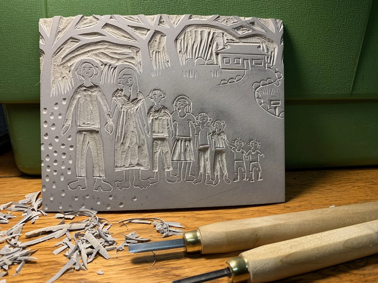 Linocut of a family holding hands outdoors, with trees and a house in the background, on a green background with carving tools and shavings in front.