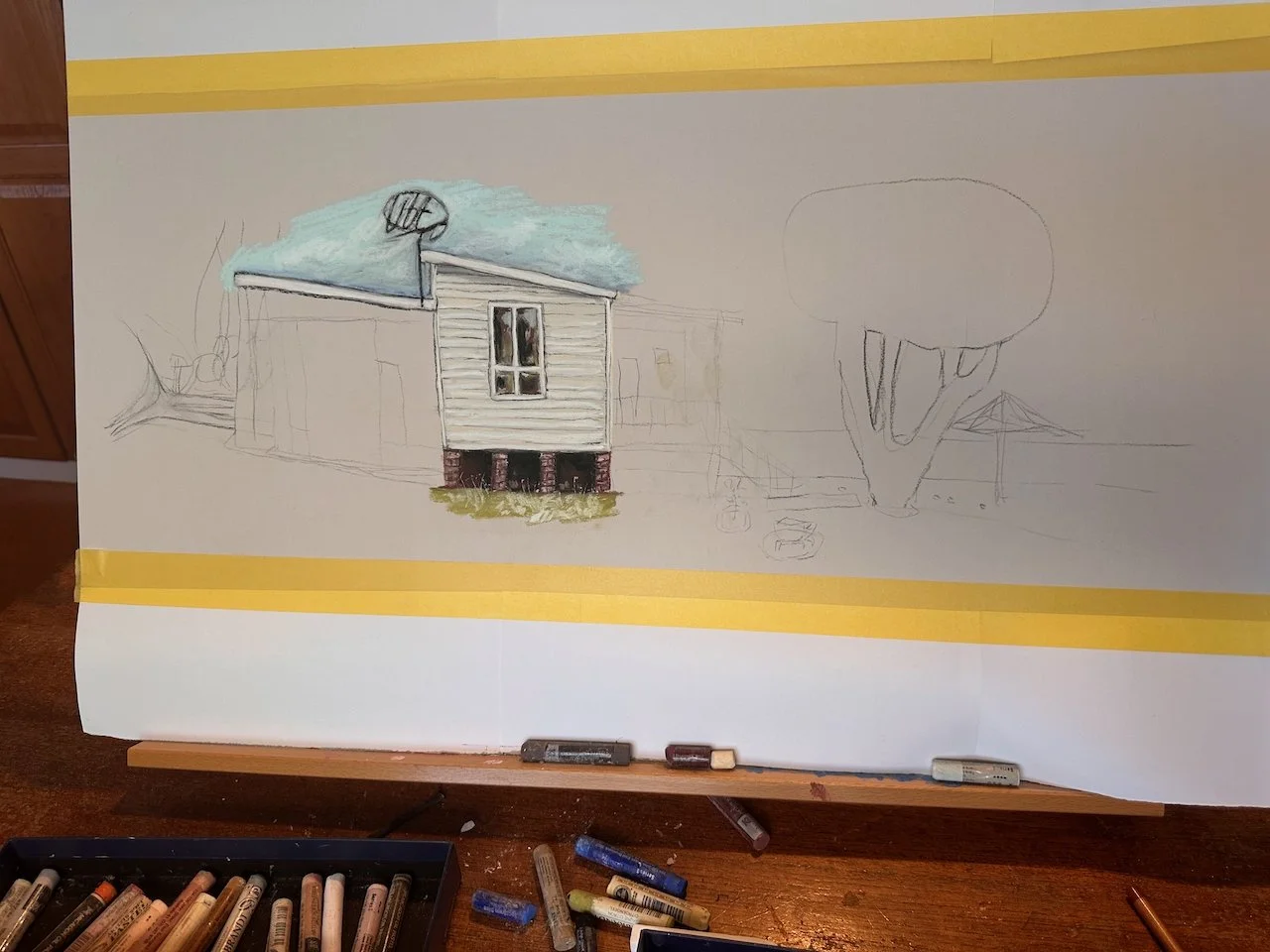 Sketch of a house with detailed front and side views, some parts colored, surrounded by other objects, and art supplies on the table.