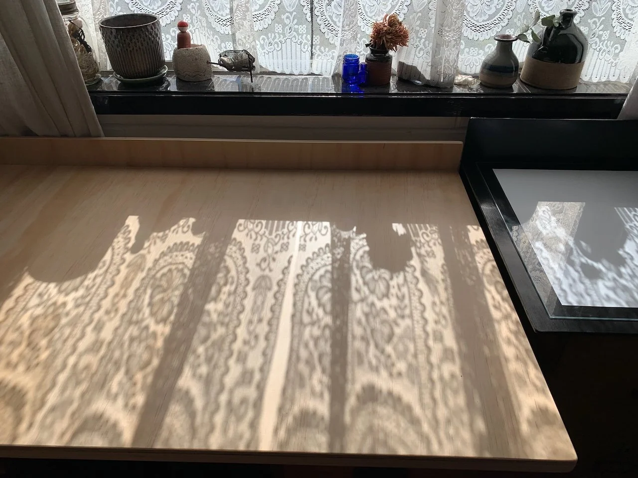 Sunlight casting intricate lace shadow patterns on a light-colored wooden table near a window with lace curtains. The window sill holds various decorative vases and containers, including a plant with dried flowers.