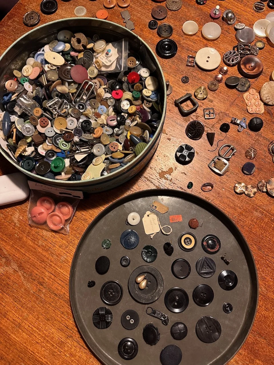 A collection of buttons of various sizes, colors, and shapes on a wooden surface. Some buttons are in a round container, and others are spread out on the table. There are also small metal fasteners, a zipper pull, and a few tags among the buttons.