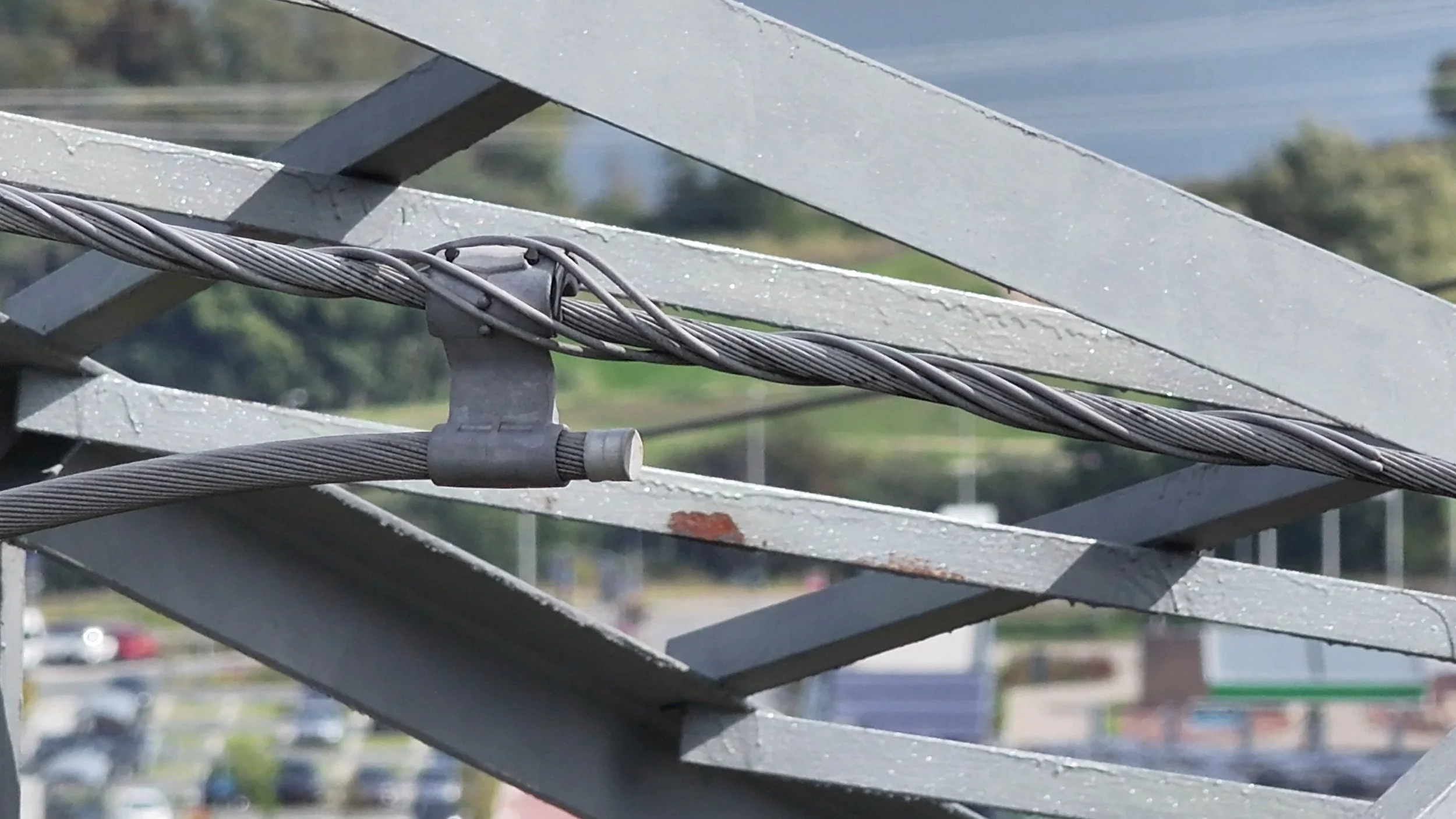 Close-up van kabels en metalen constructies op een deel van een gebouw bij zonnig weer, met op de achtergrond onscherpe parkeerplaatsen en bomen.