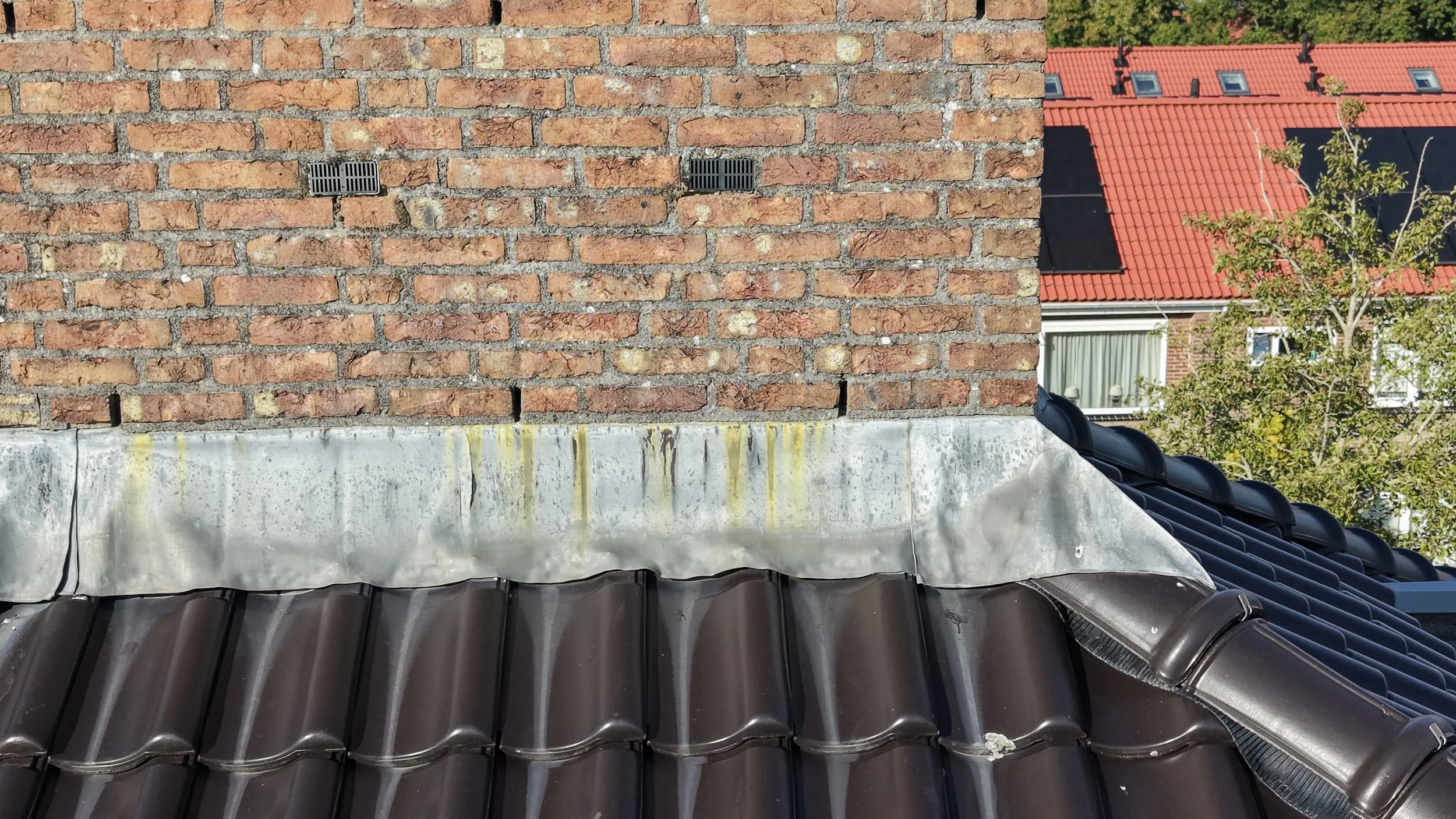 Dak met zwarte dakpannen en een gemetselde schoorsteen. Op de achtergrond staat een huis met een rood geglazuurd pannendak en meerdere kleine dakramen. Er zijn bomen en een blauwe lucht zichtbaar.