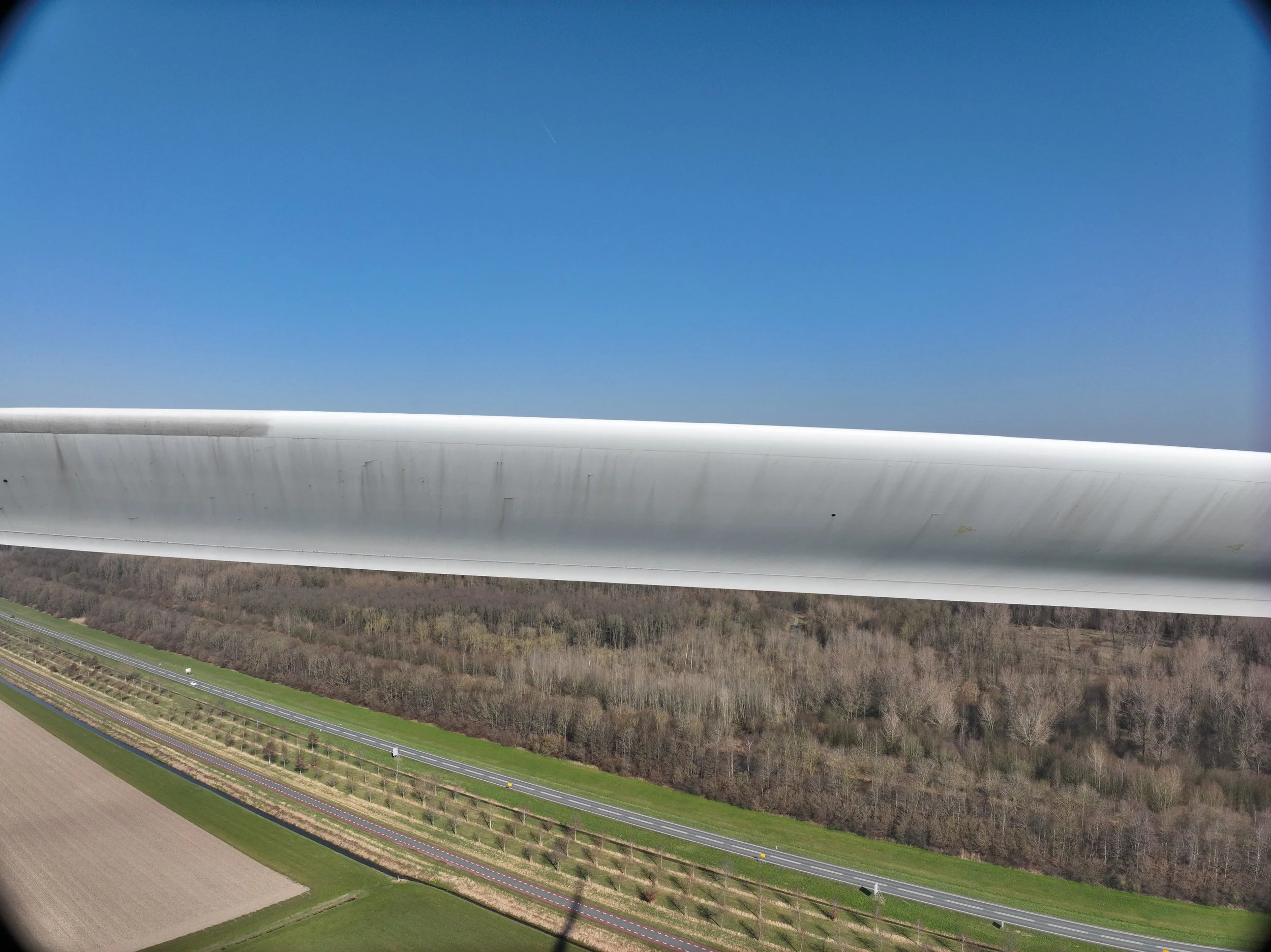 Luchtfoto van een windturbine, omgeven door landbouwvelden en bossen, onder een heldere blauwe hemel.

