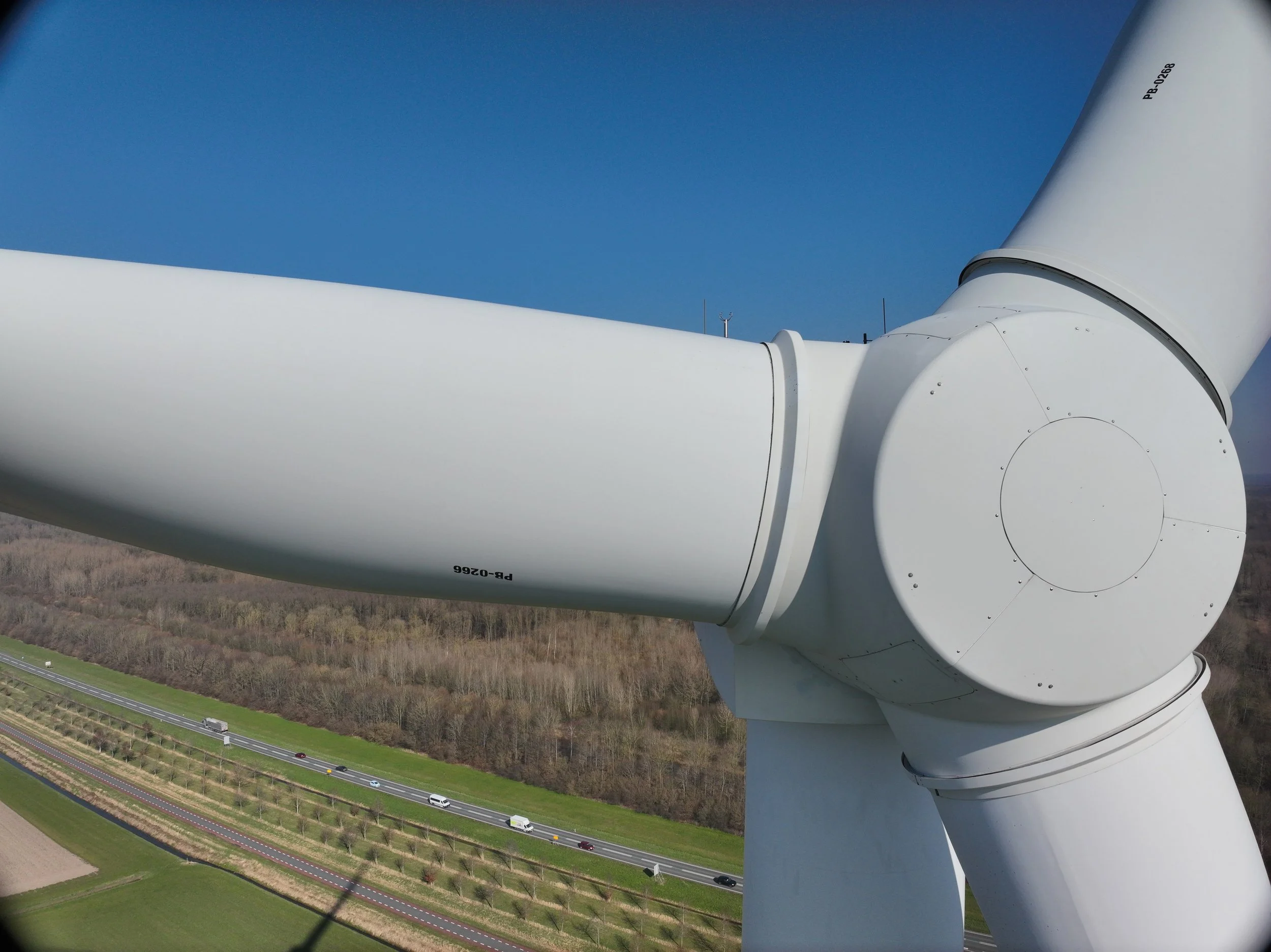 Close-up van een grote witte windturbine in de buitenlucht, met zicht op de rotor en de naaf. Op de achtergrond zijn wegen met auto’s en een landschap met velden en bomen zichtbaar.
