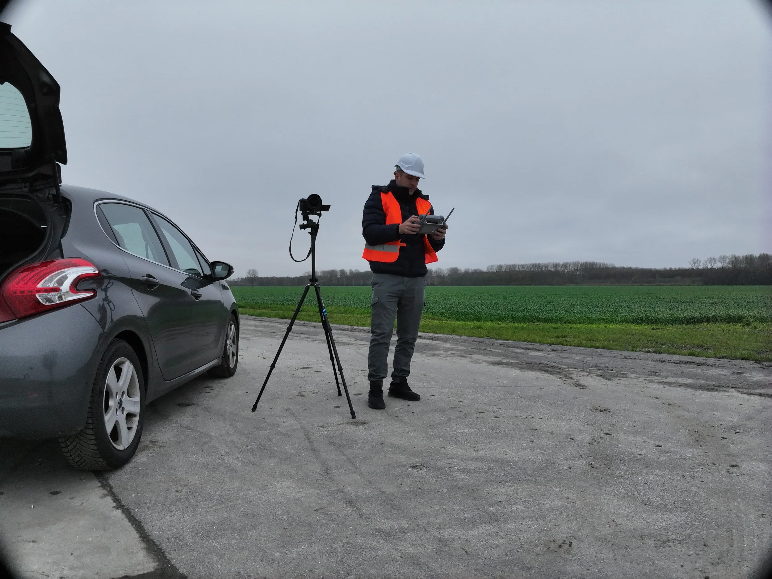 Een man met een veiligheidsvest en beschermende helm bestuurt een drone met een afstandsbediening op een open grasveld, naast een auto met een geopende kofferbak.
