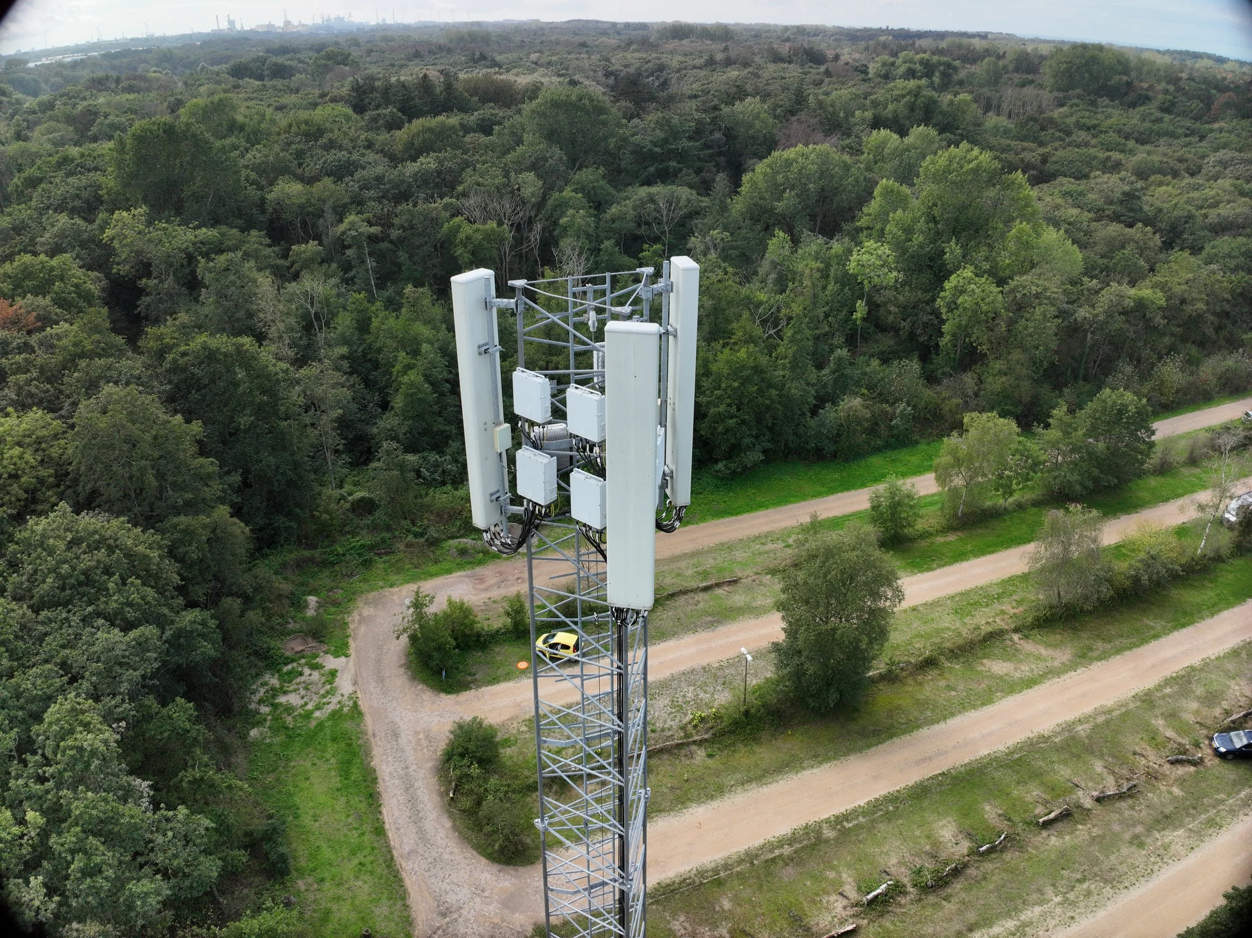 Een zendmast voor mobiele communicatie midden in een dicht bos, met een bosweg en enkele geparkeerde auto’s op de voorgrond. Op de achtergrond is een stad zichtbaar met industriële gebouwen en windturbines.