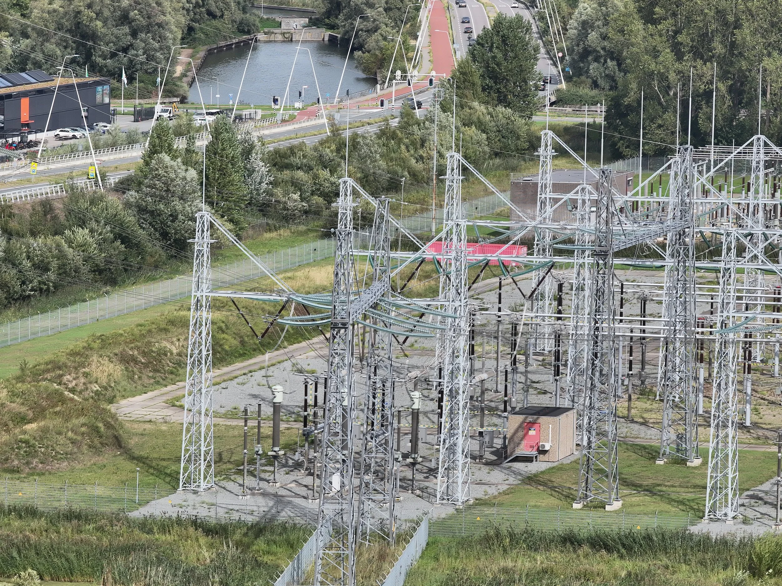Zicht op technische infrastructuur met hoogspanningsleidingen, een waterkanaal, wegen, auto’s en bomen in een stedelijke omgeving.