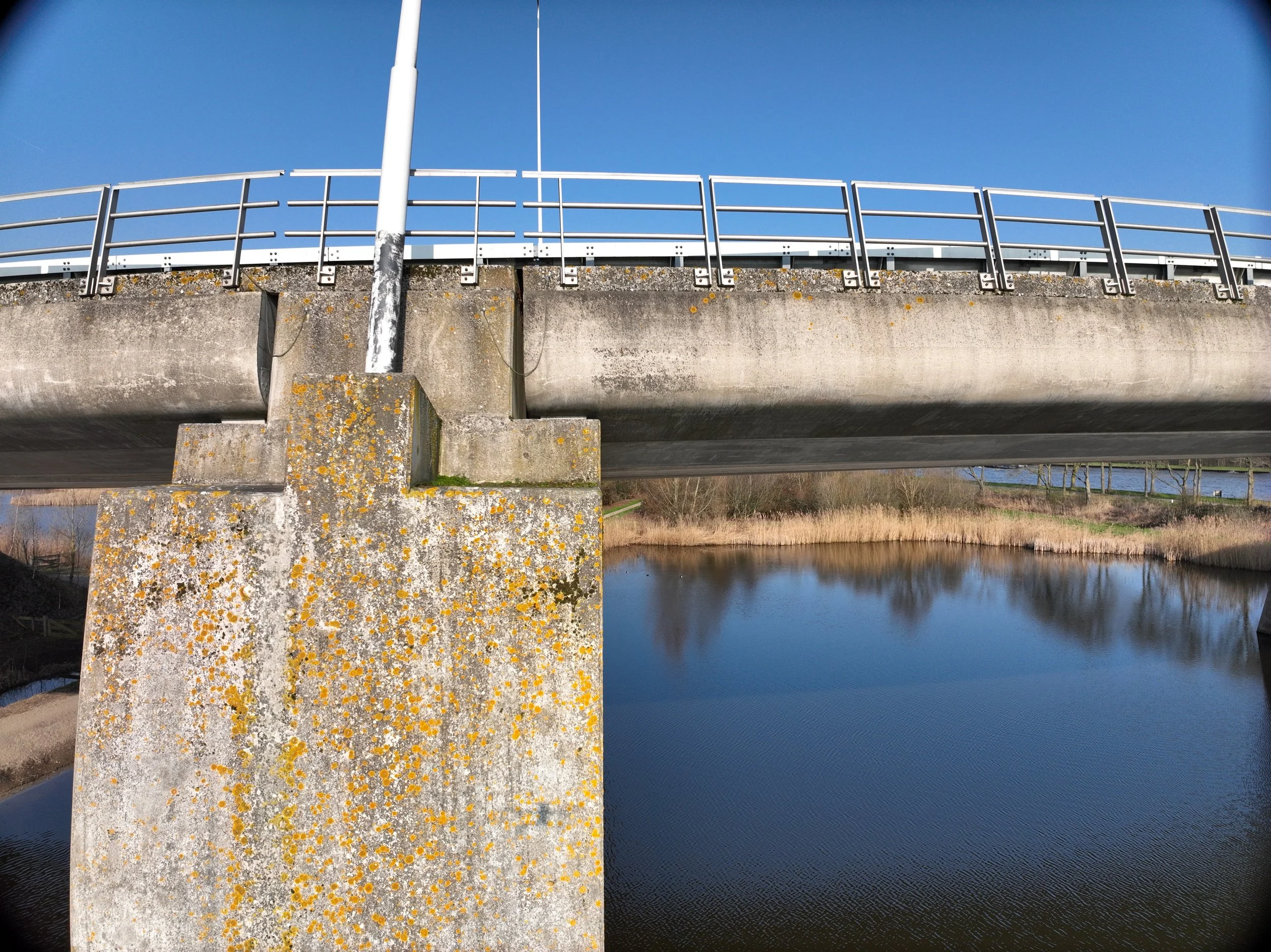 Brug over een rivier onder een zonnige hemel, met betonnen pijlers en metalen vangrails. Langs het water bevindt zich een groene oever.