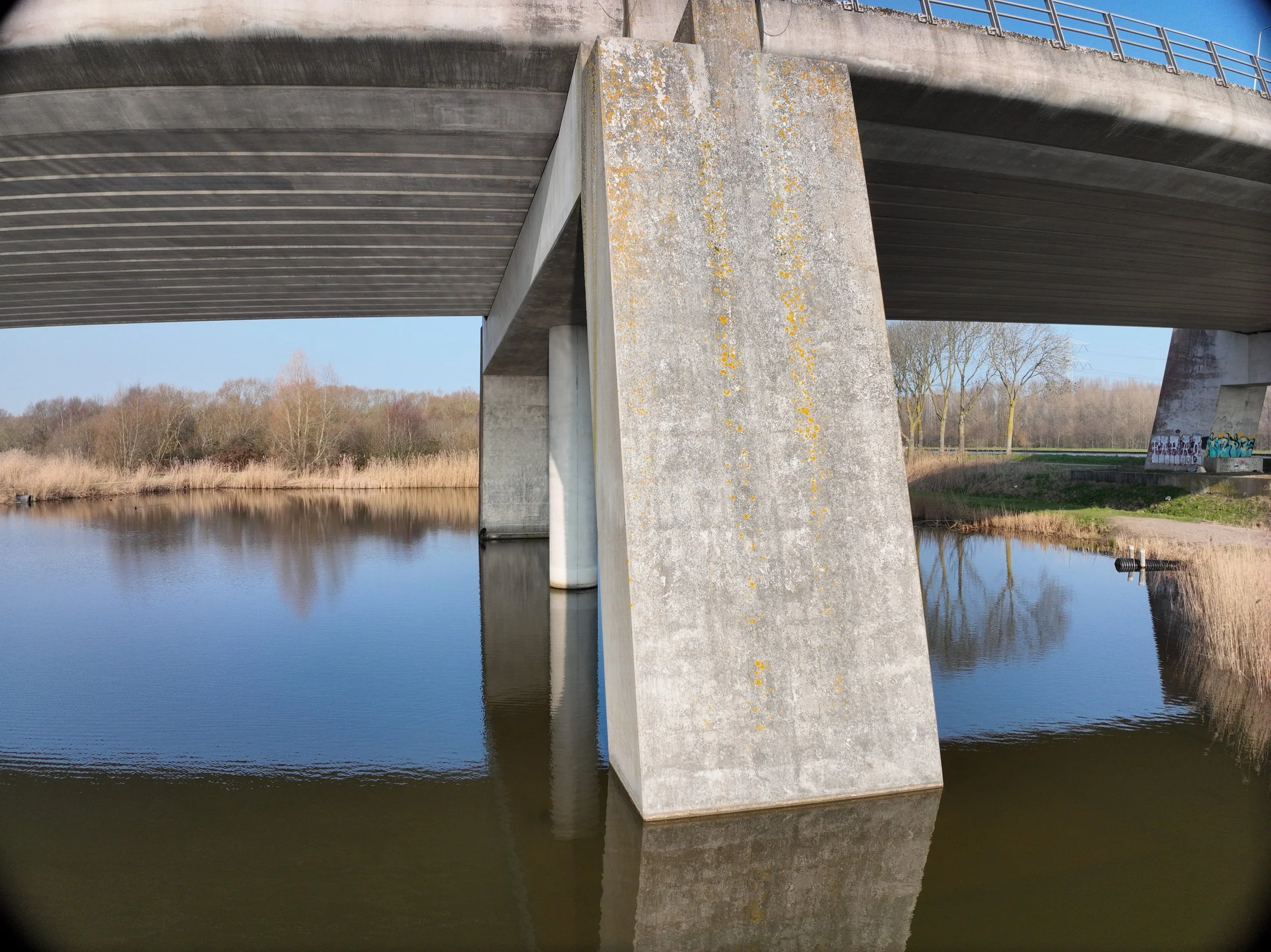 Zicht onder een brug over een rivier, met een grote betonnen constructie en een reling langs de brug. De rivier weerspiegelt de lucht en de constructie, met bomen langs de oever op de achtergrond.