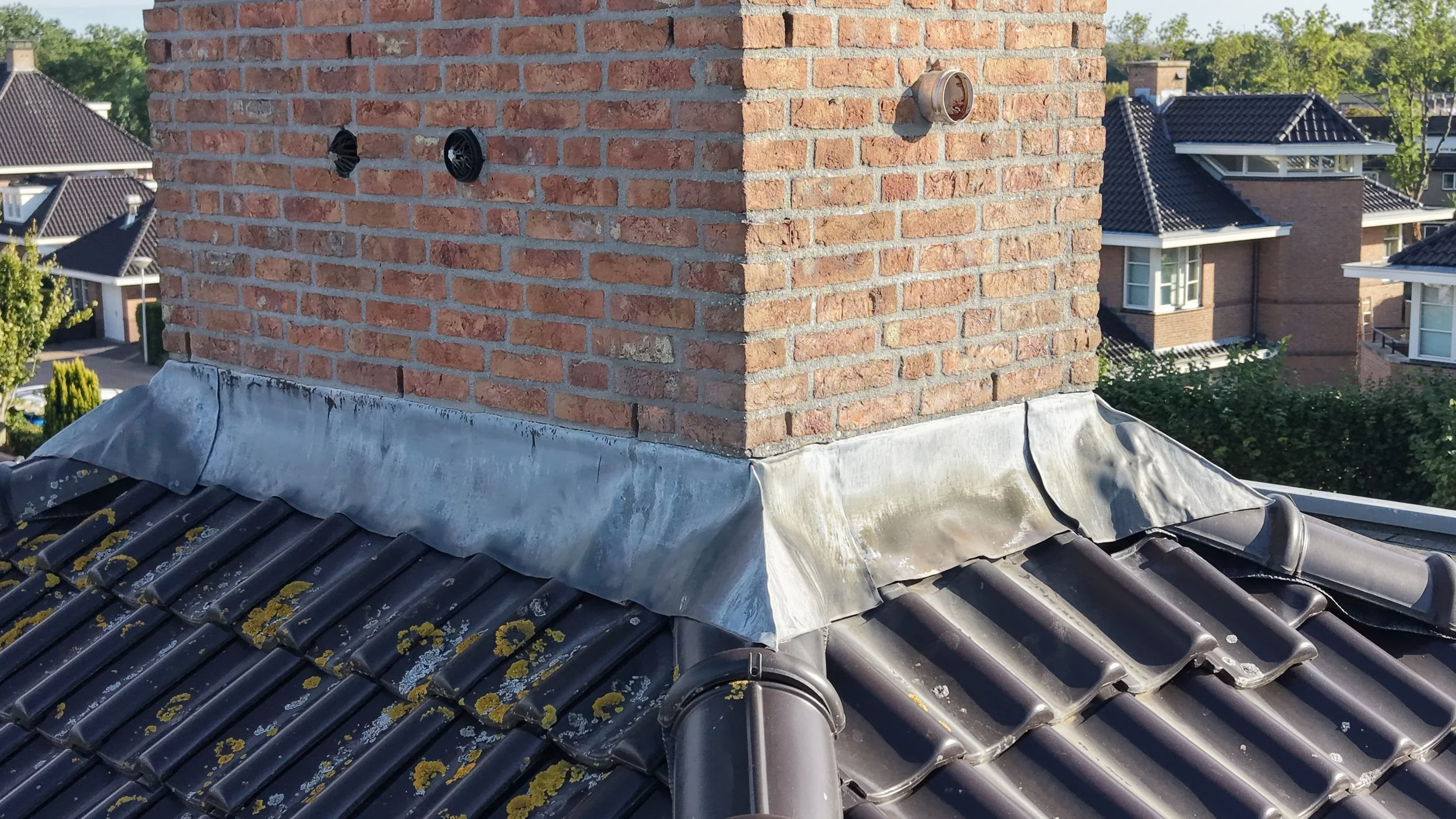 Zicht op een dakdoorvoer met schoorsteen op een groen dak. Op de voorgrond zijn dakpannen en zilverfolie zichtbaar, op de achtergrond staan woonhuizen met donkere daken.