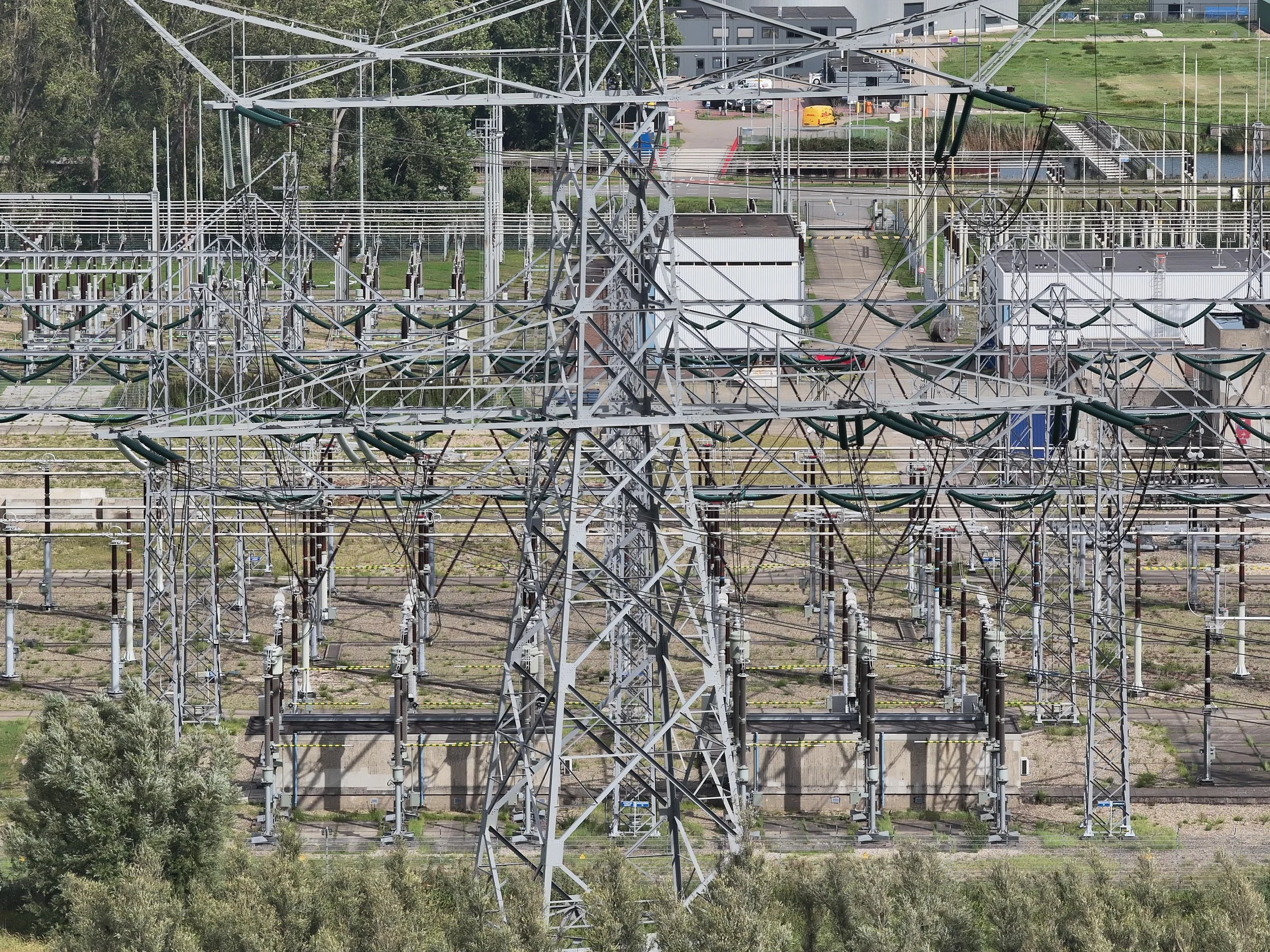 Een elektriciteitsmast op een elektriciteitsstation met meerdere kabels en leidingen, omgeven door groene bomen en een buiteninstallatie met gebouwen en een weg op de achtergrond.