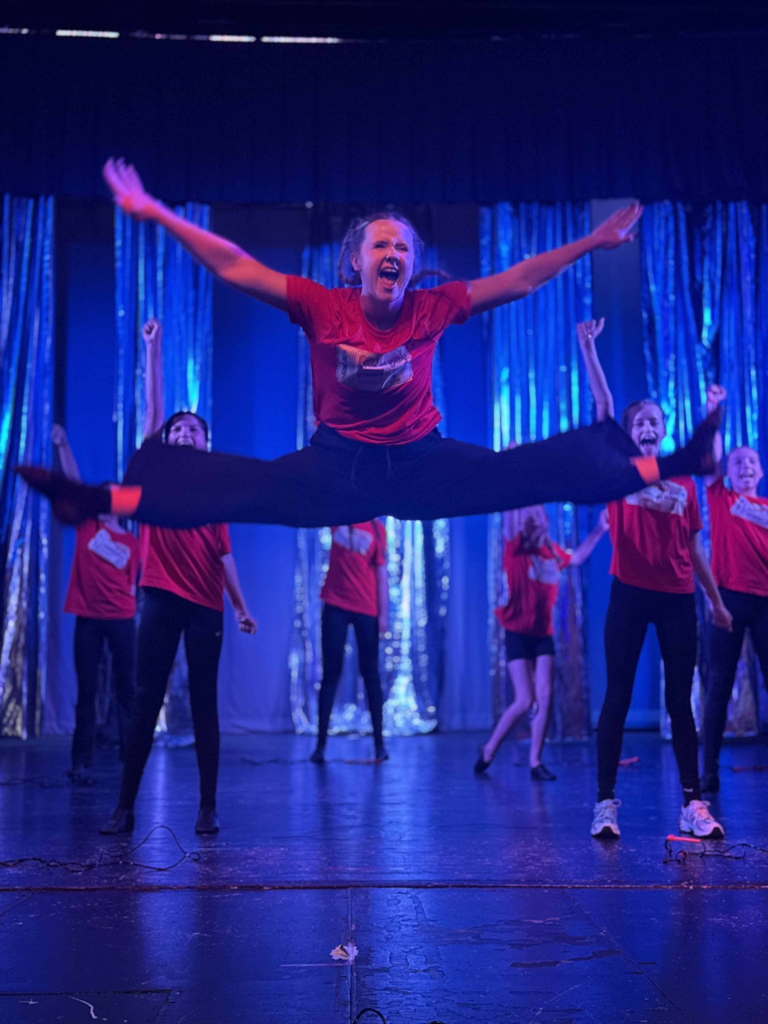 Group of performers on stage in a dance routine, with a woman in a red shirt mid-jump with legs spread wide and arms outstretched, with others in red shirts behind her, blue stage lights, and a shiny backdrop.
