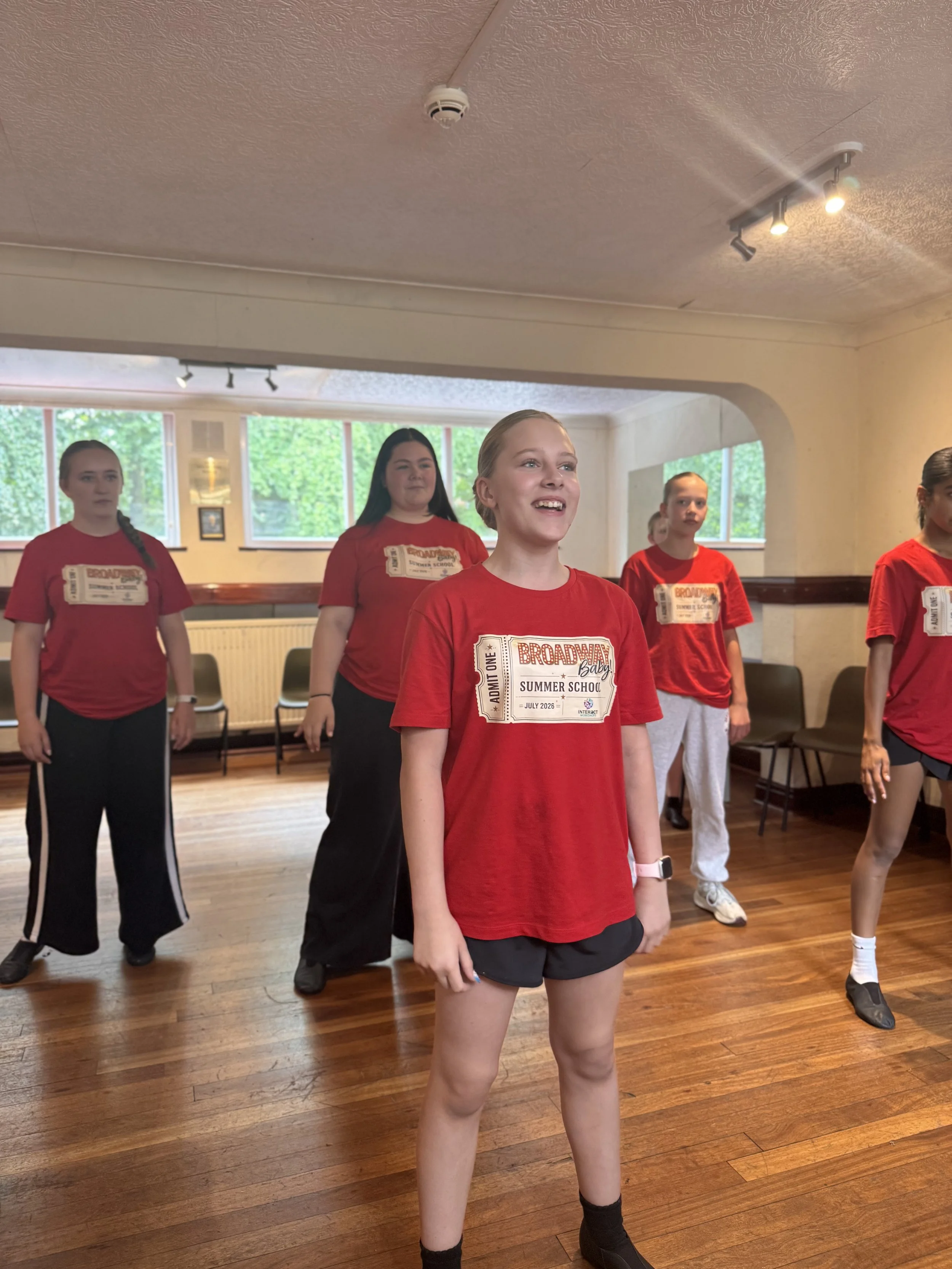 Group of young girls participating in a dance class in a studio, standing on a wooden floor, wearing matching red t-shirts with a summer school logo, some with their arms at their sides and others with hands on their hips.